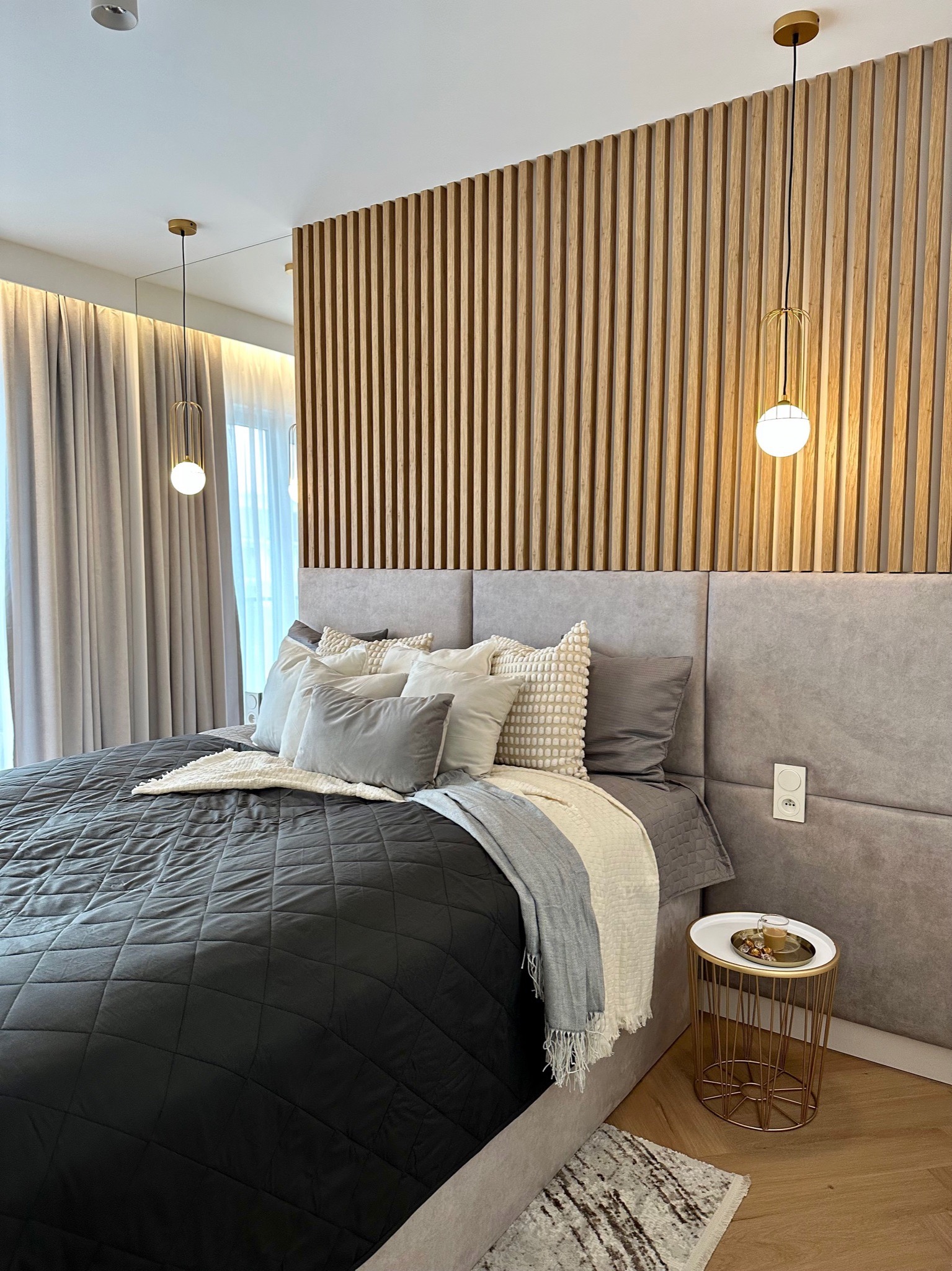 Nowoczesna sypialnia z drewnianą ścianą z lameli, szarą tapicerowaną ramą łóżka, czarną pikowaną narzutą, dekoracyjnymi poduszkami, stolikiem nocnym z kawą i wiszącymi lampami z kulistymi kloszami.