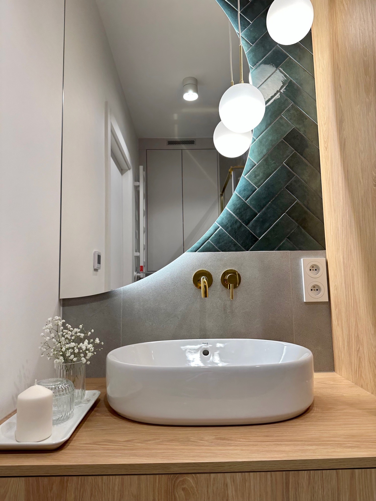 Nowoczesna łazienka z owalną umywalką nablatową, złotą armaturą, lustrem o nieregularnym kształcie i zielonymi płytkami ułożonymi w jodełkę, dopełniona dekoracyjną świecą i kwiatami.