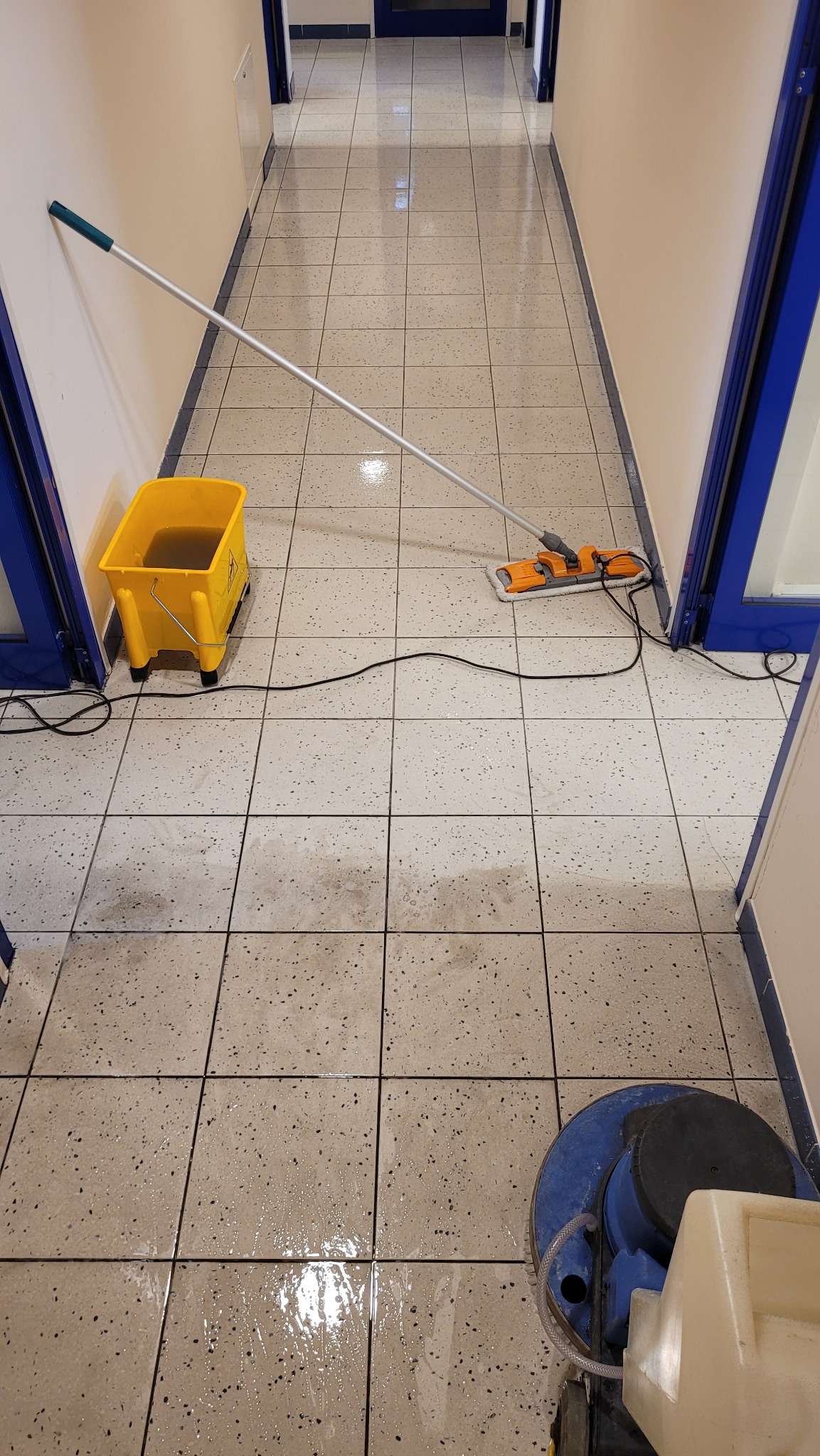 Korytarz z terakotą w trakcie czyszczenia: mop parowy z żółtym wiadrem i automat szorujący do podłóg w akcji.