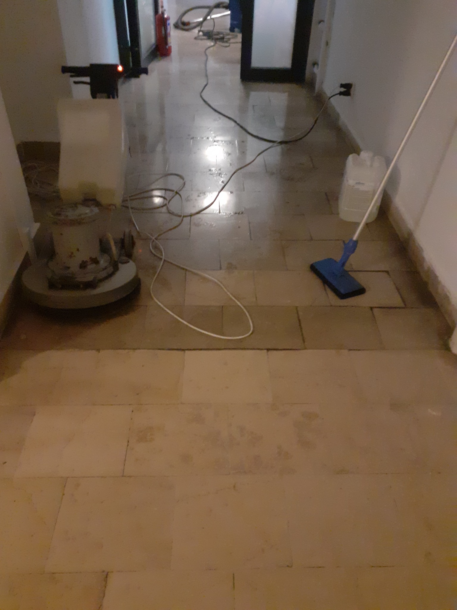 Polerka do podłóg i mop na mokro na korytarzu z kamienną posadzką w trakcie czyszczenia, widoczny także kanister z płynem i przewody zasilające