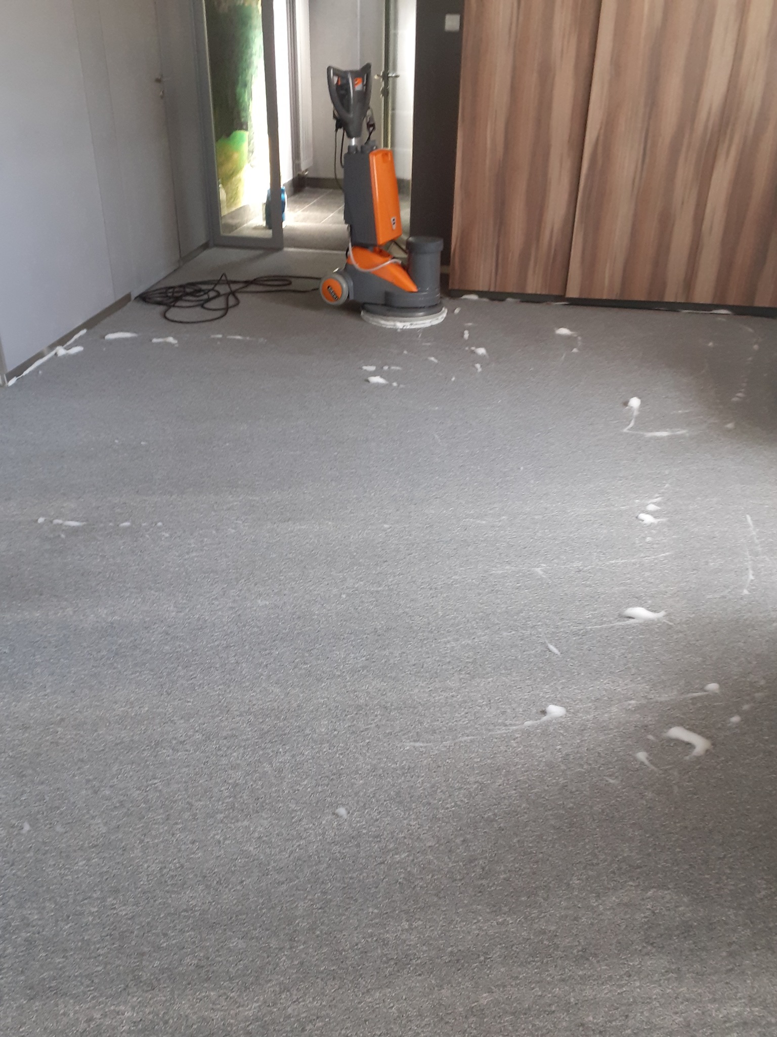 Maszyna do czyszczenia podłóg w trakcie pracy na szarym dywanie, widoczne ślady piany i świeżo wyczyszczona powierzchnia.