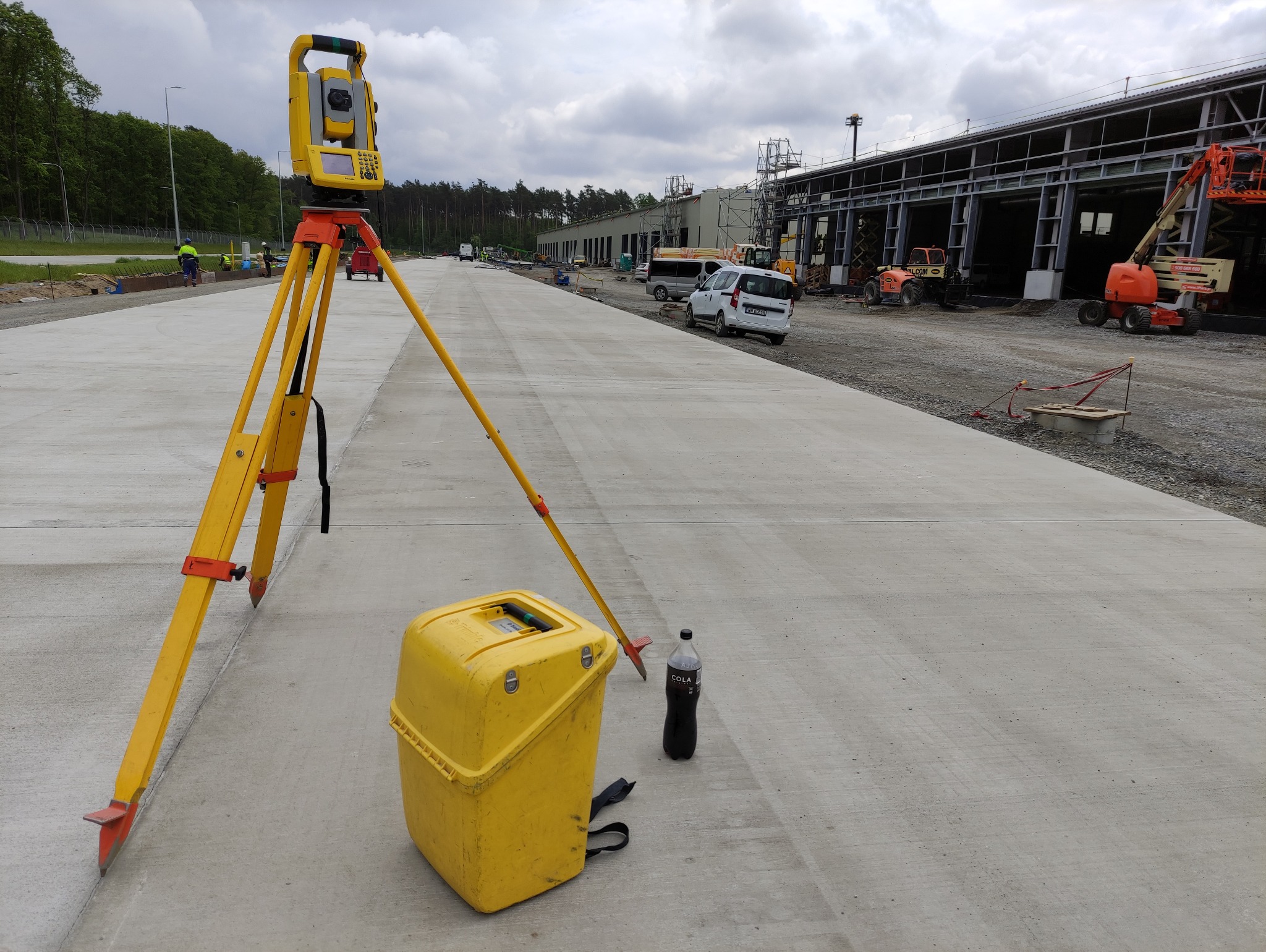 Żółty teodolit ustawiony na betonowej nawierzchni placu budowy, obok żółta skrzynka narzędziowa i butelka coli, w tle hala w budowie i sprzęt budowlany.