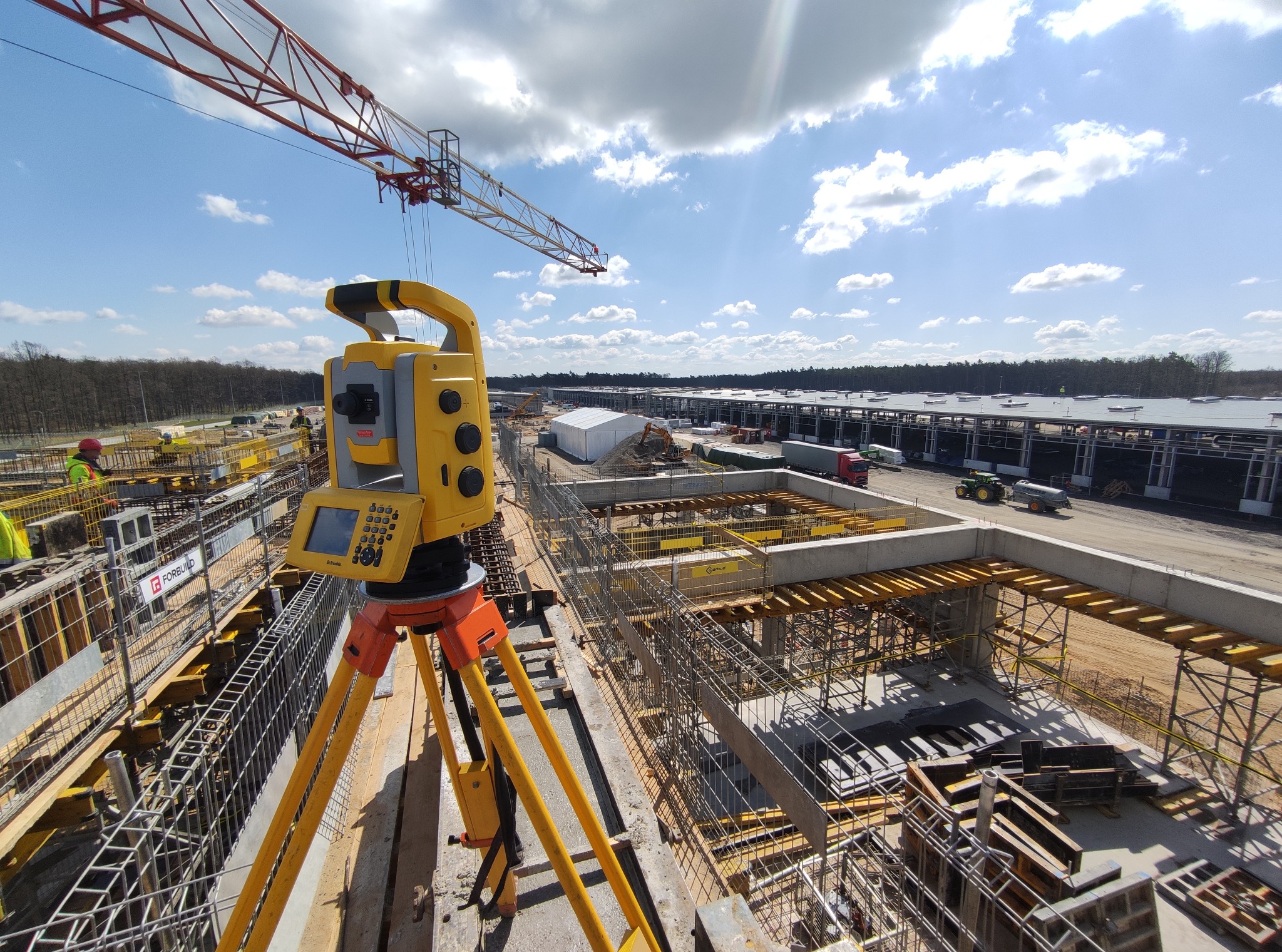 Żółty teodolit na statywie na tle budowy hali, widoczny dźwig i konstrukcja zbrojeniowa, słoneczny dzień.