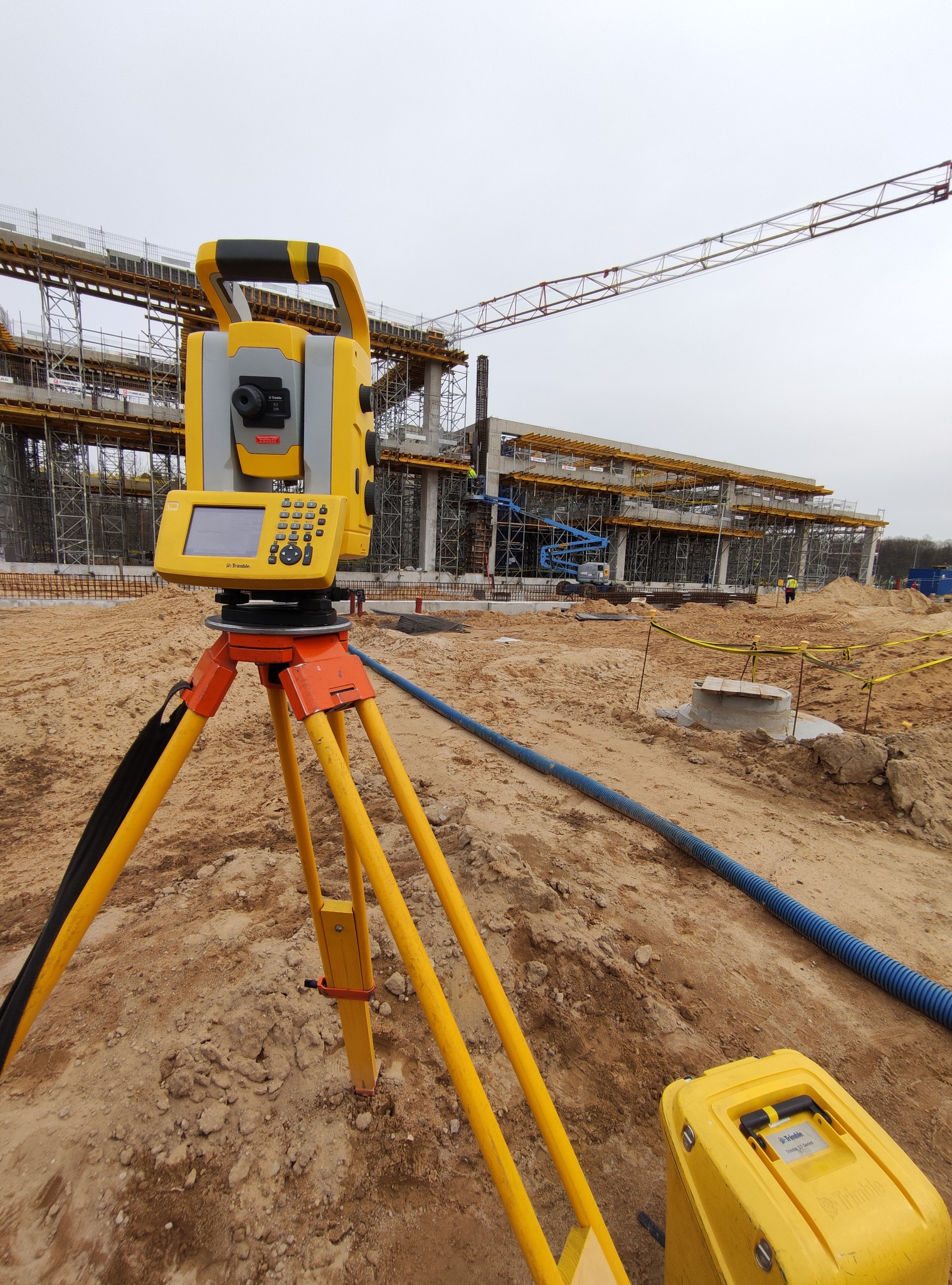 Żółty teodolit marki Trimble ustawiony na trójnogu na placu budowy, w tle konstrukcja budynku z widocznym dźwigiem i robotnikami.