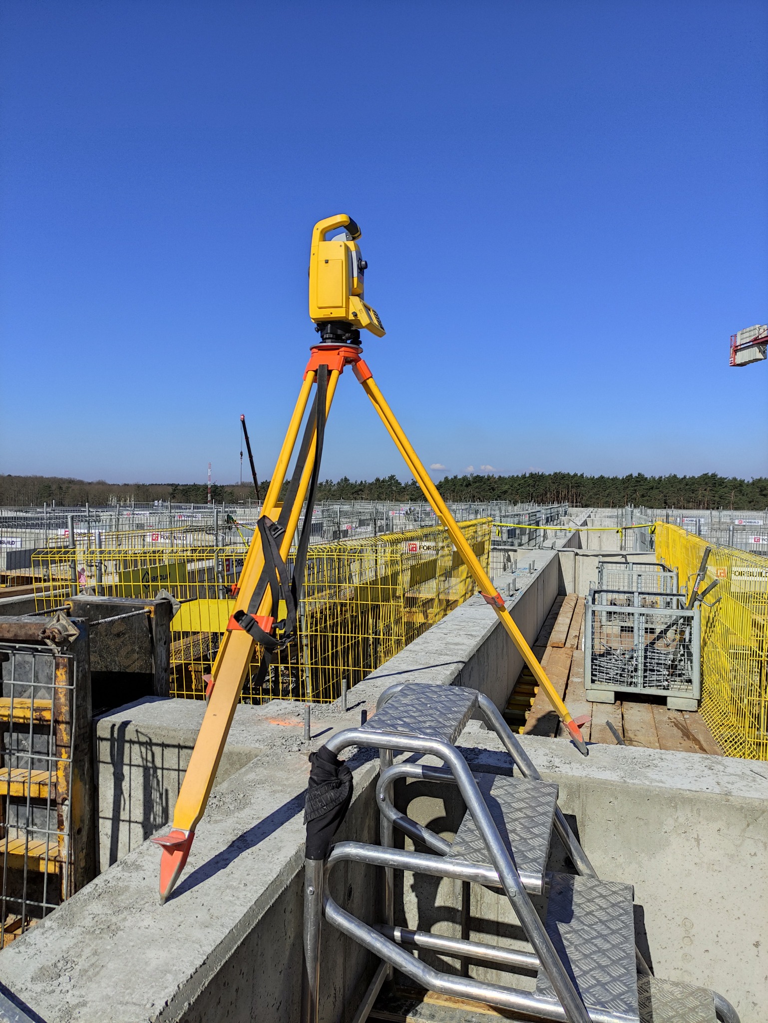 Żółty teodolit ustawiony na statywie na terenie budowy, widoczne szalunki i schody metalowe, w tle las pod błękitnym niebem.