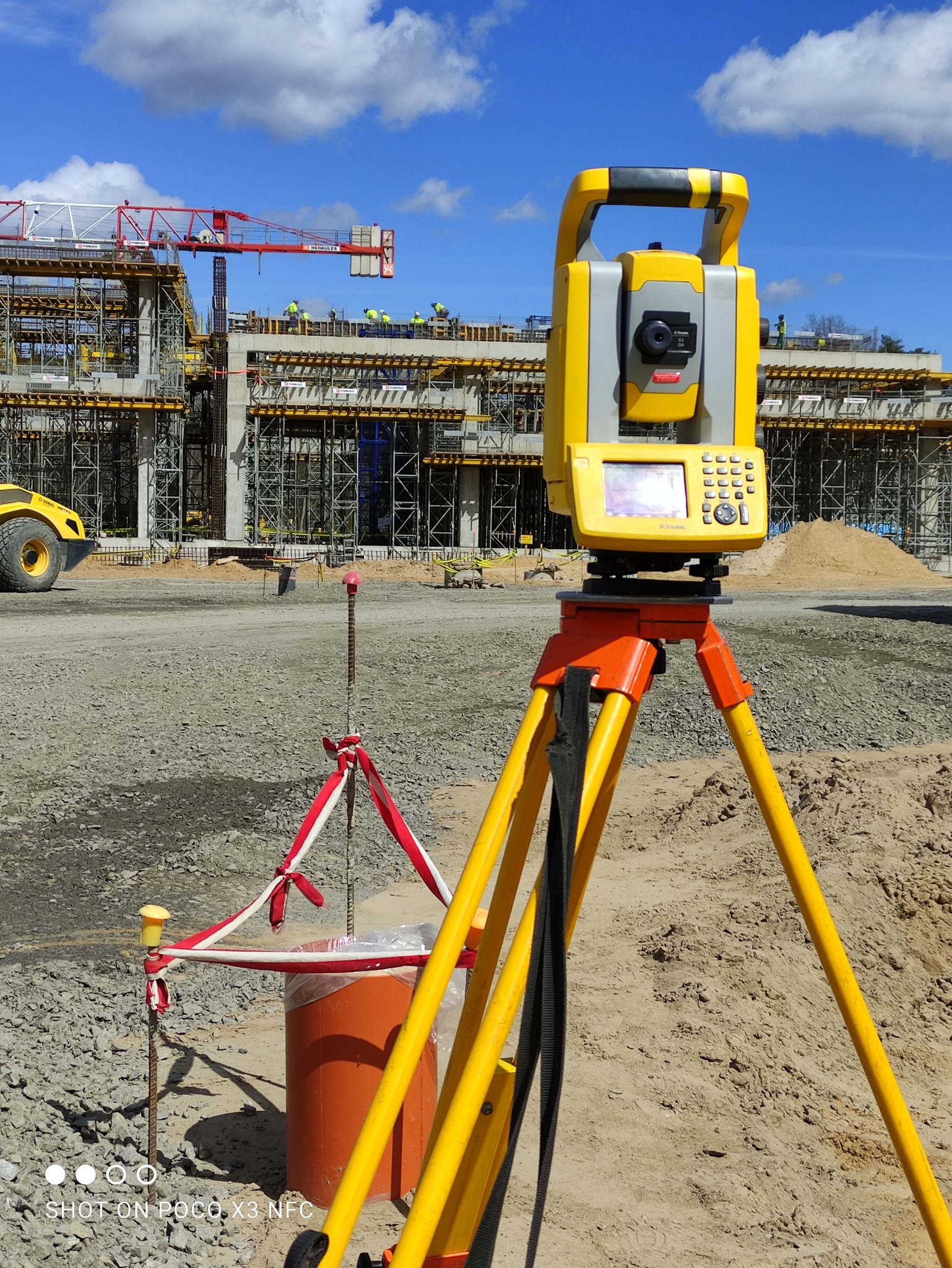 Żółty teodolit na pomarańczowym statywie na terenie budowy, w tle konstrukcja budynku z dźwigiem i robotnikami.