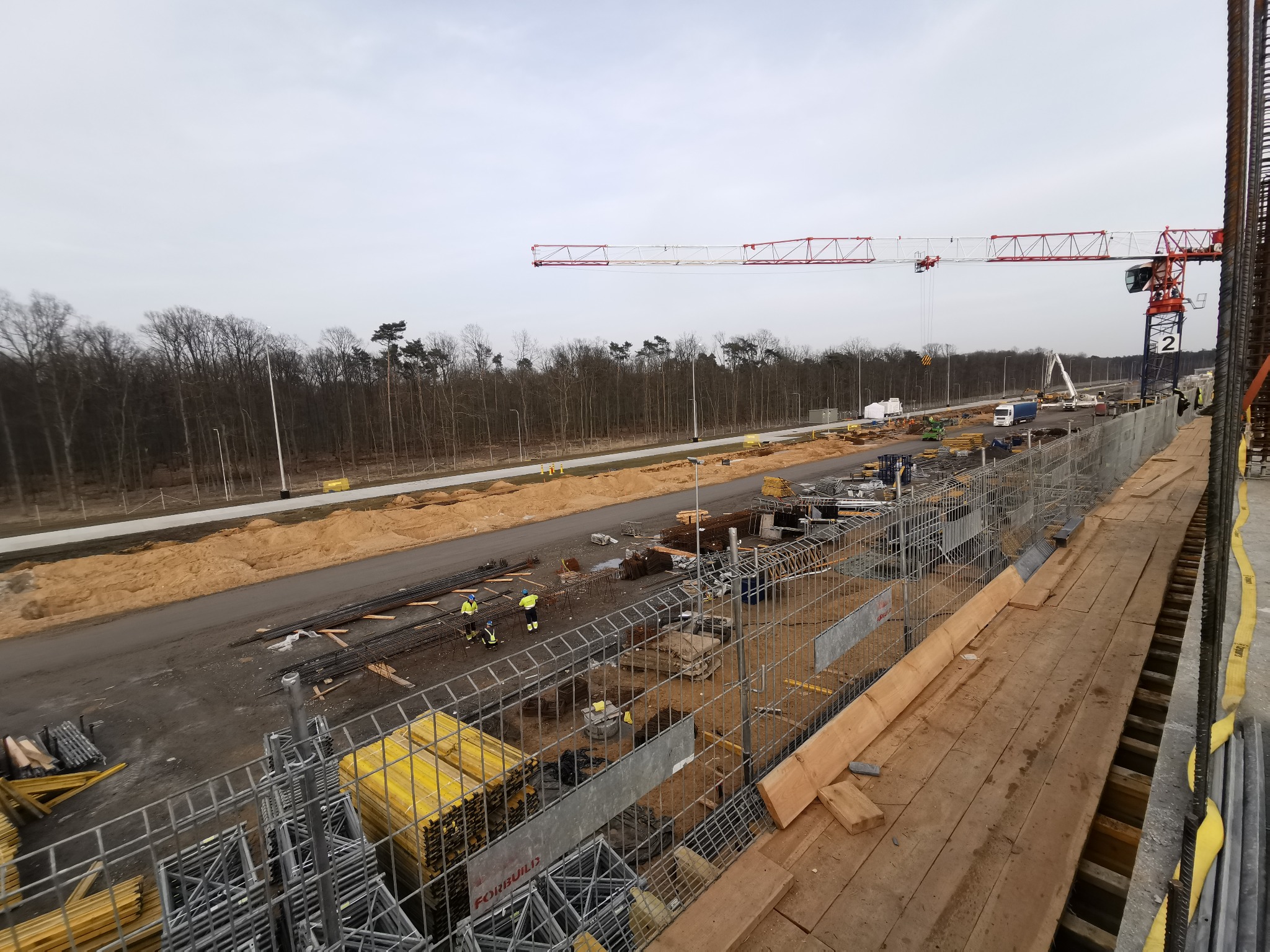 Budowa drogi z widocznymi pracownikami w odblaskowych kamizelkach, dźwigiem w tle i elementami konstrukcyjnymi na pierwszym planie, wykonana w pochmurny dzień.