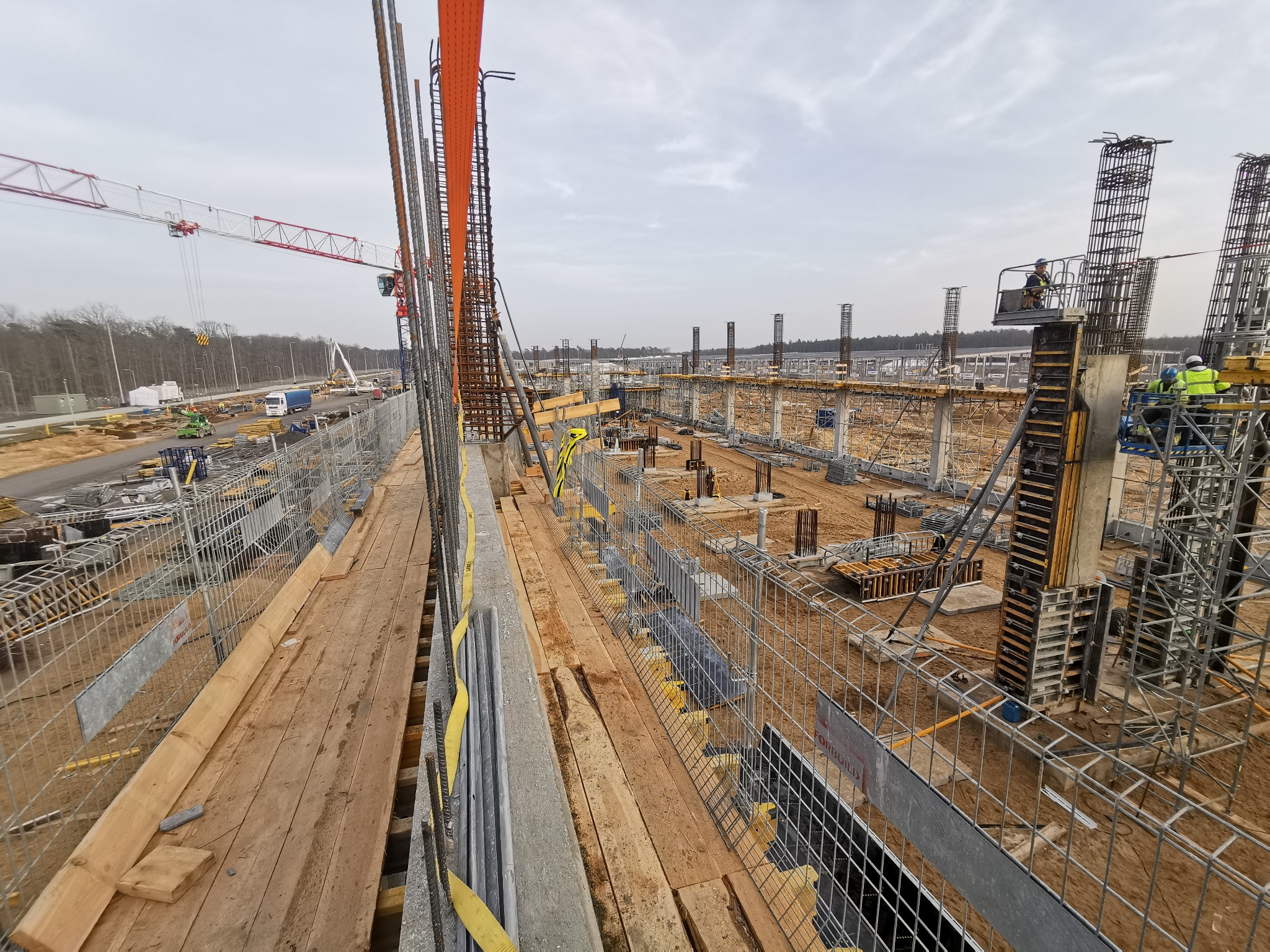 Budowa hali: widok z góry na plac budowy z widocznymi fundamentami, szalunkami, zbrojeniami, rusztowaniami i pracującymi robotnikami w oddali, oraz żurawiem w tle.