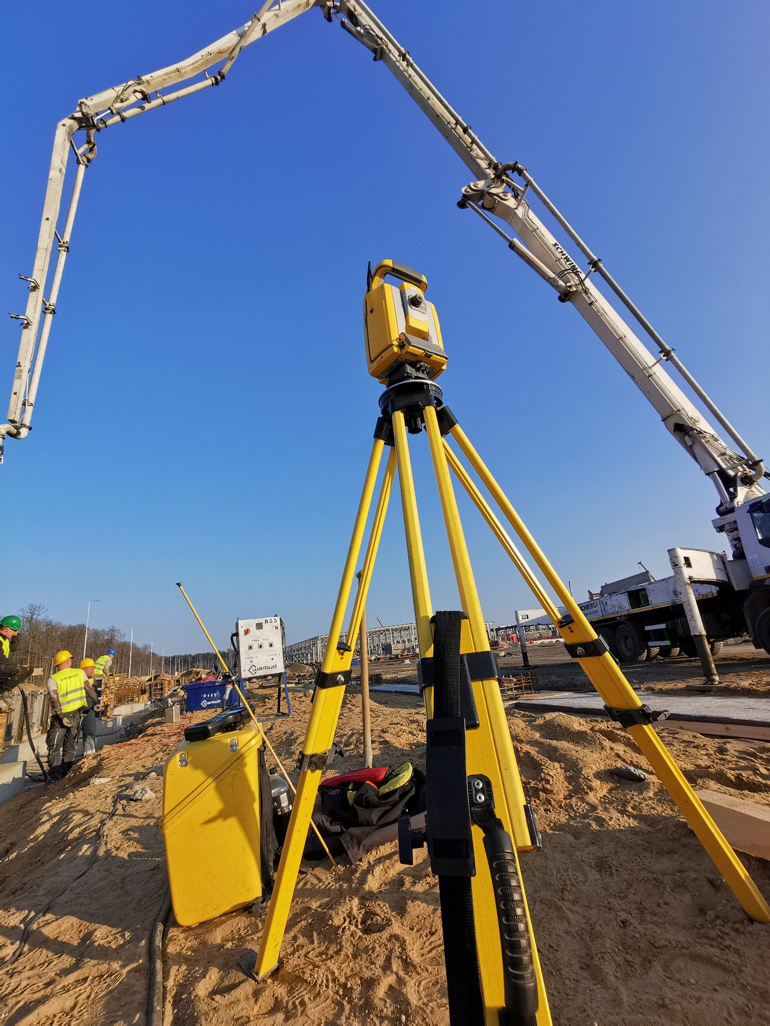 Praca geodezyjna w Rychwale: żółty teodolit na trójnogu na placu budowy, w tle pracownicy w kaskach i betonopompa Schwing.