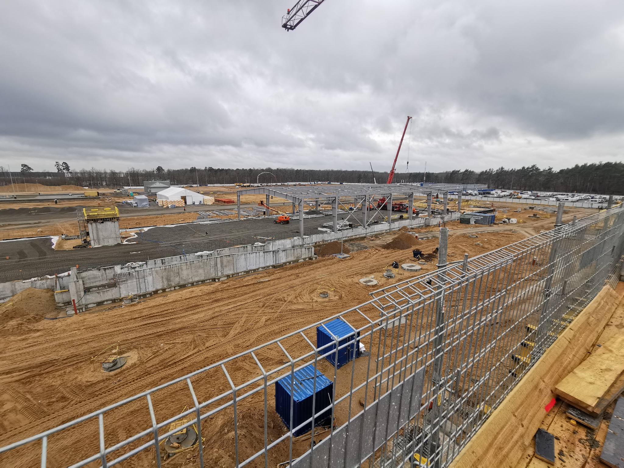 Szeroki kadr na plac budowy z konstrukcją stalową, widoczne fundamenty, dźwigi i kontenerowe biura, w tle las i pochmurne niebo.