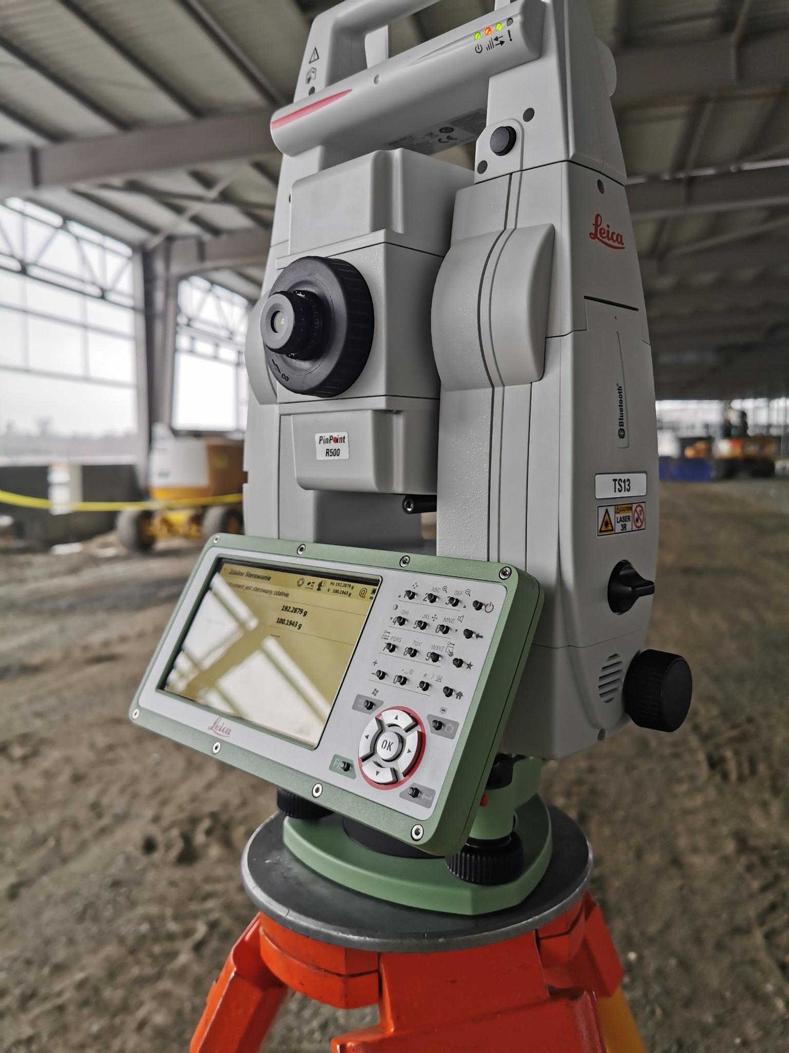 Profesjonalny teodolit Leica TS13 ustawiony na trójnogu, z widocznym ekranem i panelem sterowania, na tle placu budowy z żółtą taśmą ostrzegawczą i maszyną budowlaną w tle.