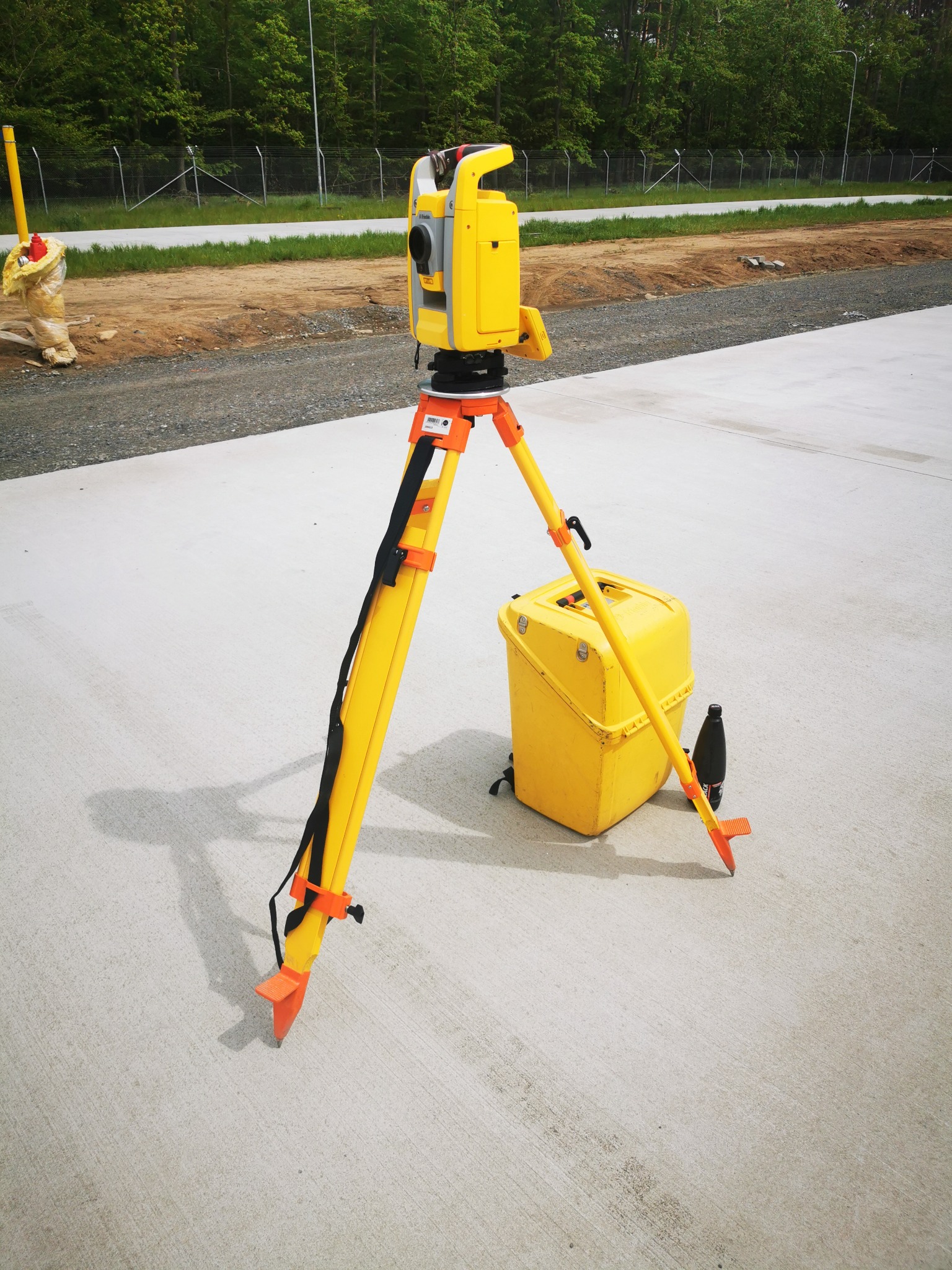 Żółty teodolit na trójnogu stoi na betonowej powierzchni, obok żółta skrzynka, w tle teren budowy z siatką ogrodzeniową i drzewami.