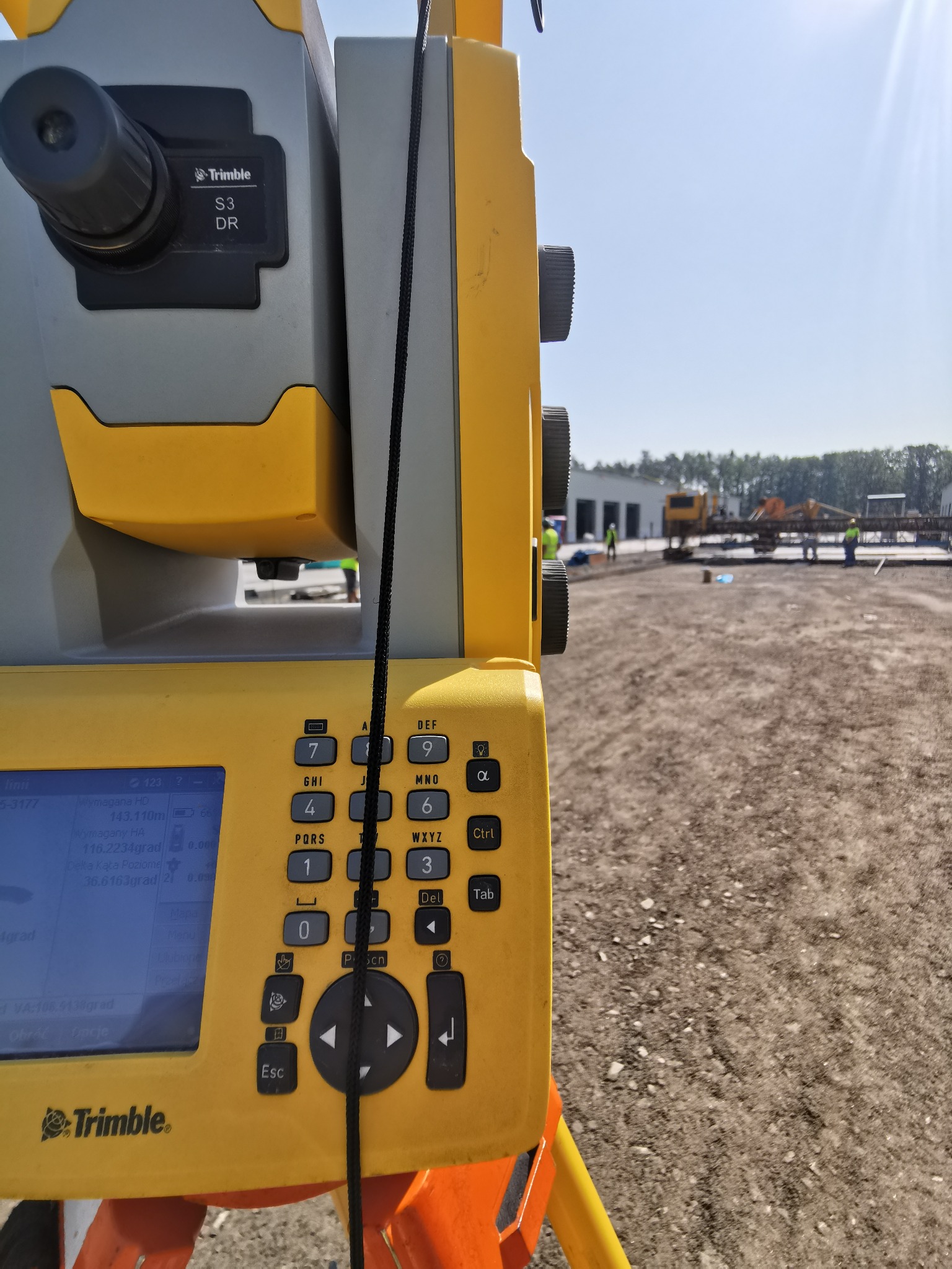 Żółto-szary teodolit Trimble S3 DR ustawiony na placu budowy z widocznymi pracownikami i sprzętem w tle, ekran urządzenia wyświetla dane pomiarowe.