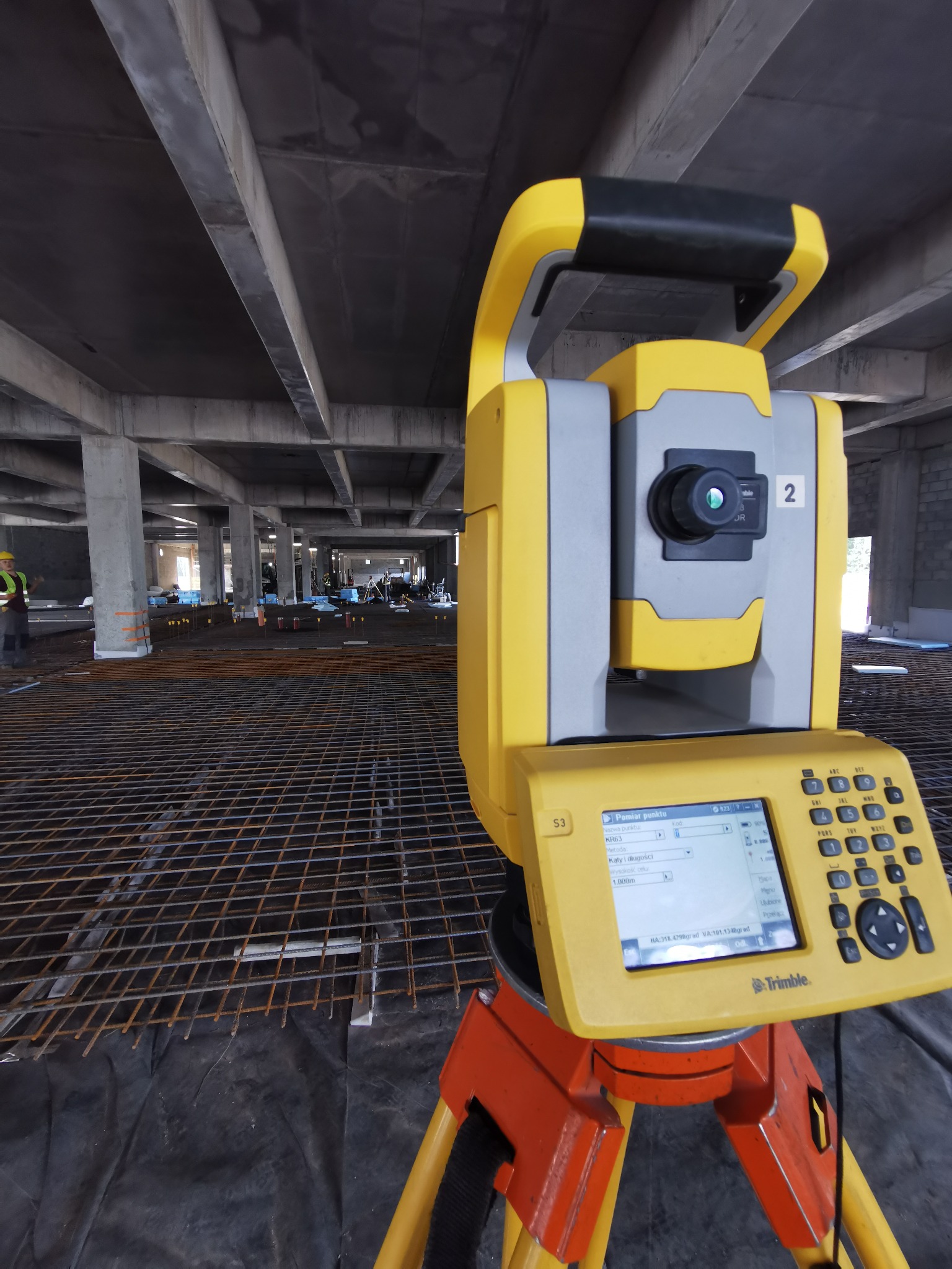Żółto-szary tachimetr Trimble na placu budowy, ustawiony na tle zbrojenia stropu i betonowych słupów, widoczny pracownik w oddali.