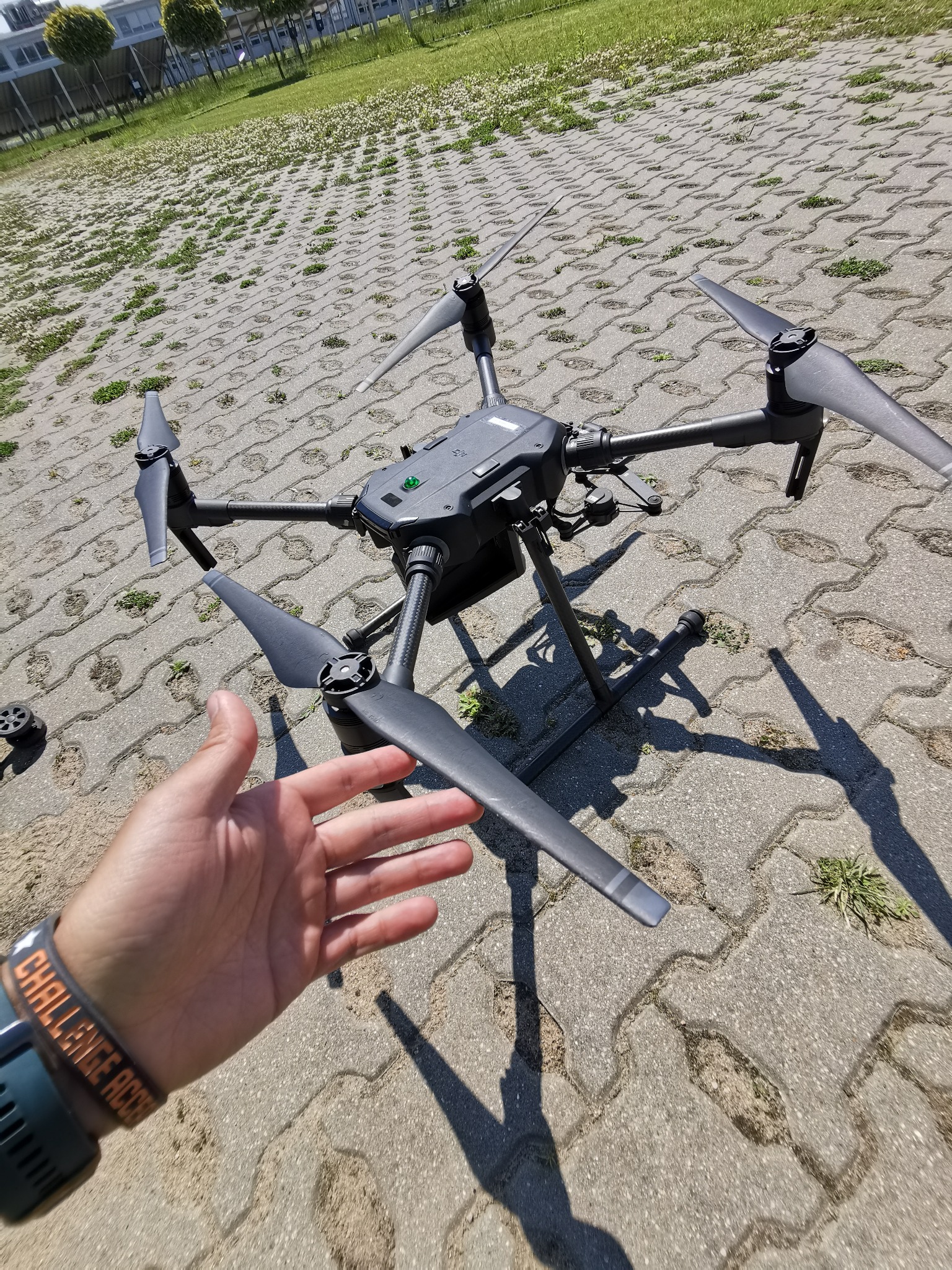 Czarny dron na rozłożonym podwoziu, dłoń z zegarkiem z napisem 'CHALLENGE ACCEP', kostka brukowa w tle.
