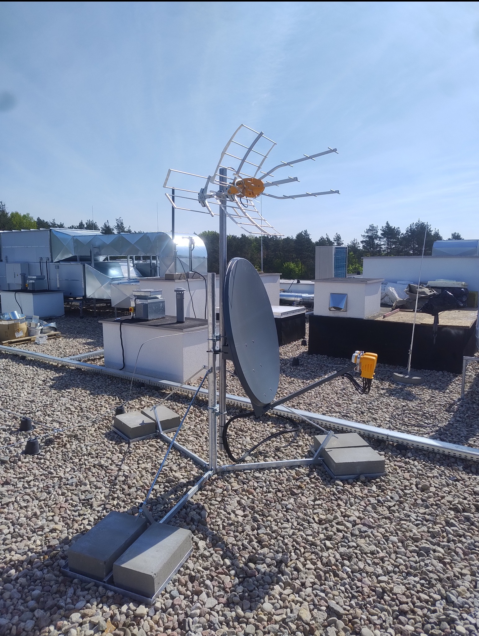 Antena satelitarna i naziemna na dachu budynku z widocznymi elementami instalacji i wentylacji. Słoneczny dzień, błękitne niebo.