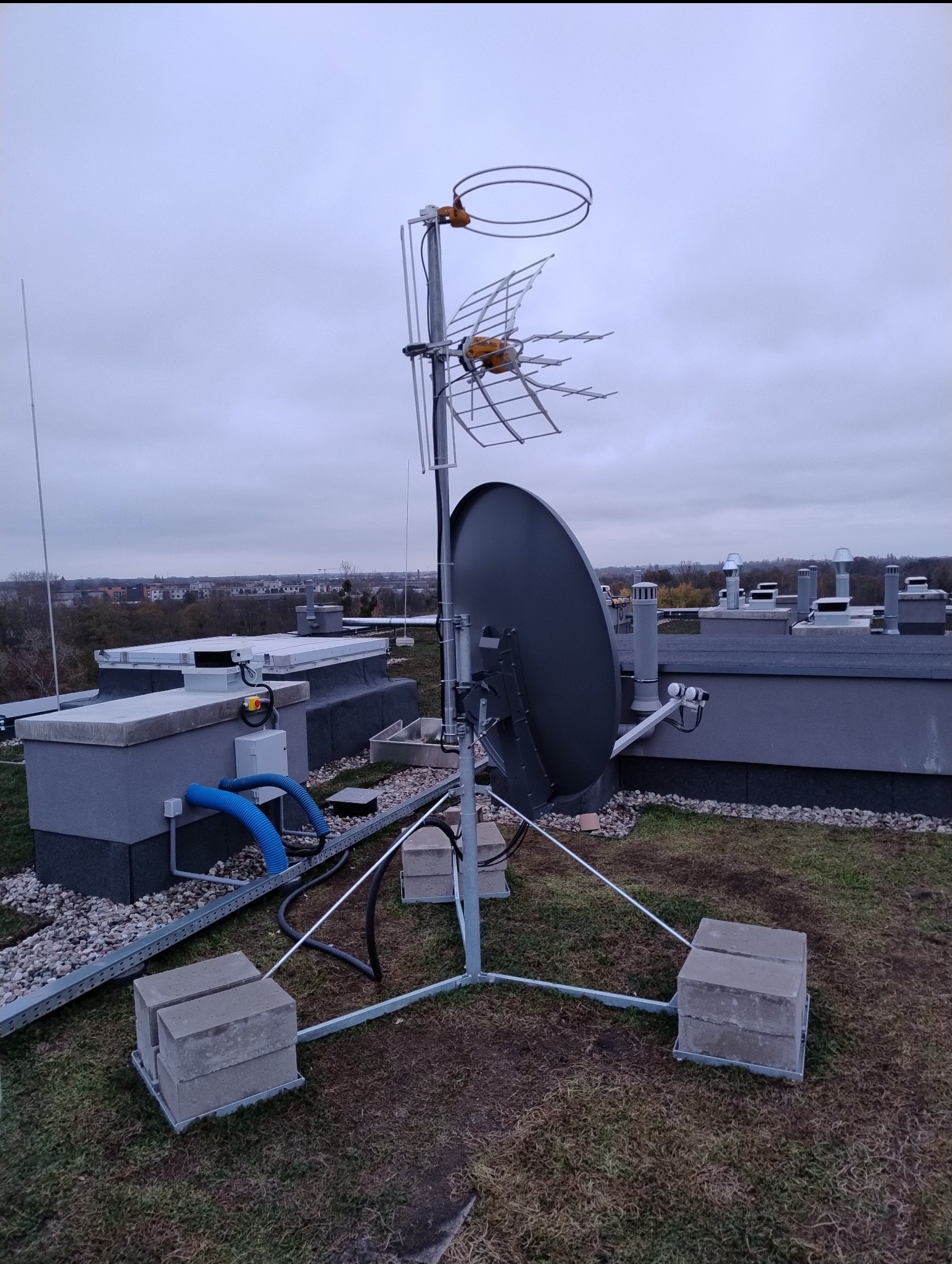 Antena satelitarna i telewizyjna na dachu budynku, zamontowana na betonowych blokach. Widoczne przewody i elementy instalacji. W tle zabudowa miejska.
