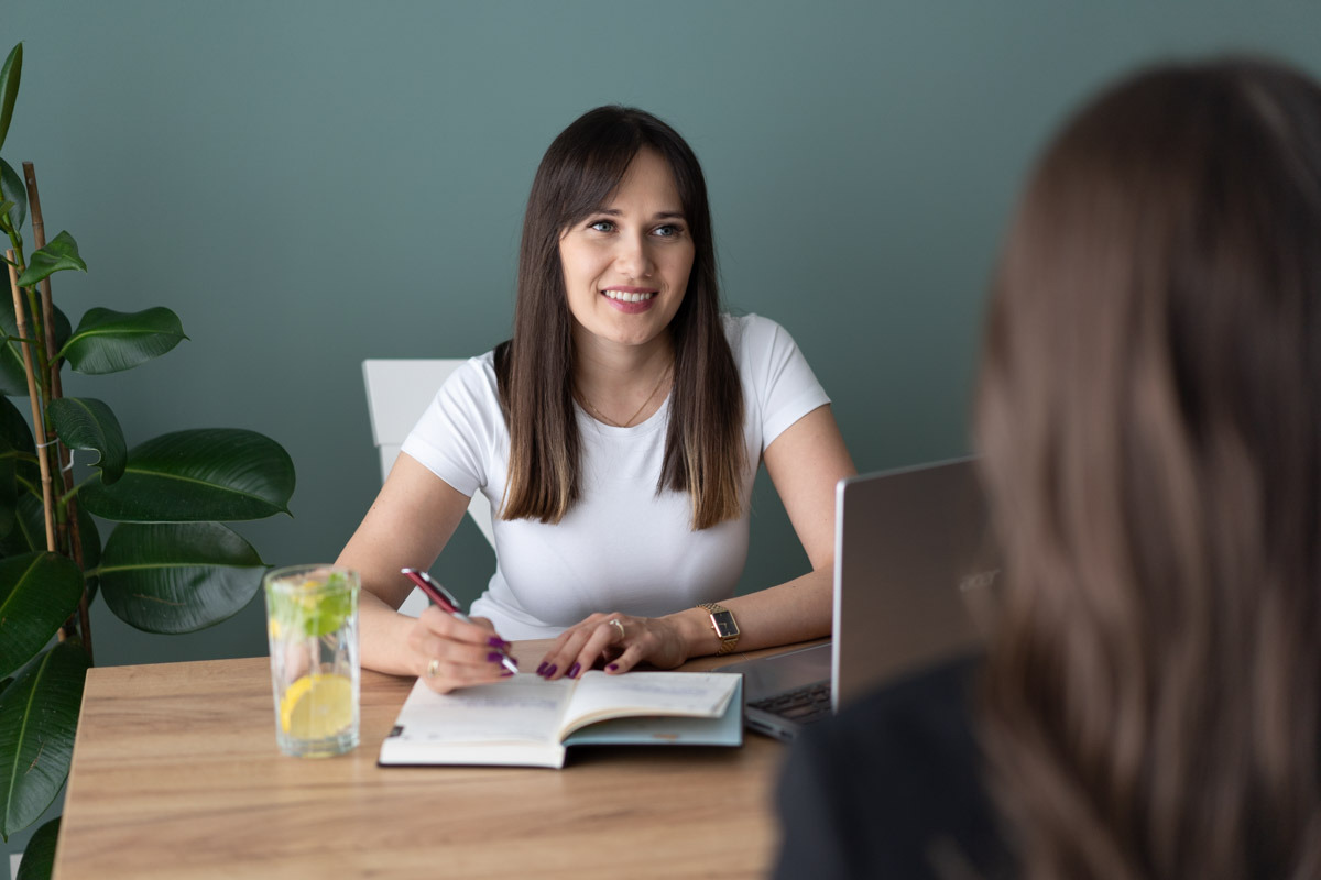 Uśmiechnięta kobieta z długimi brązowymi włosami zapisuje coś w otwartym notatniku na drewnianym biurku podczas konsultacji. Na biurku szklanka wody z cytryną i miętą, laptop oraz roślina...