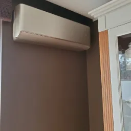 Nowoczesny klimatyzator ścienny w kolorze szampańskim zamontowany w rogu pokoju z brązowymi ścianami, obok białej stolarki okiennej.