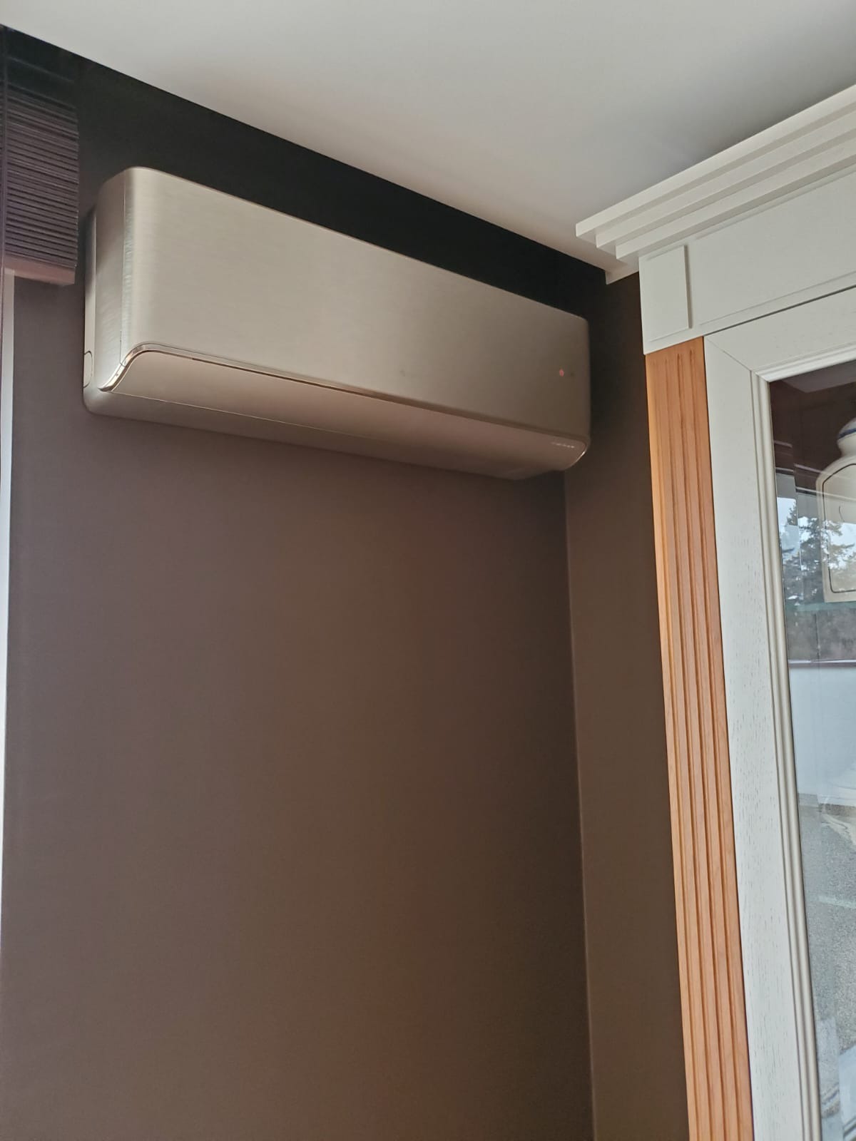 Nowoczesny klimatyzator ścienny w kolorze szampańskim zamontowany w rogu pokoju z brązowymi ścianami, obok białej stolarki okiennej.