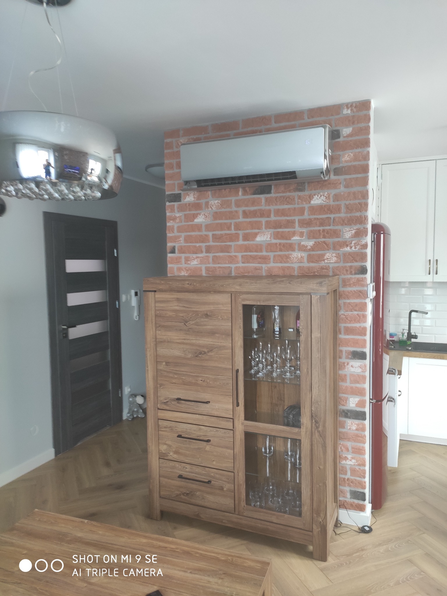 Nowoczesny klimatyzator ścienny zamontowany nad drewnianą komodą na tle ściany z czerwonej cegły w jasnym wnętrzu mieszkania.