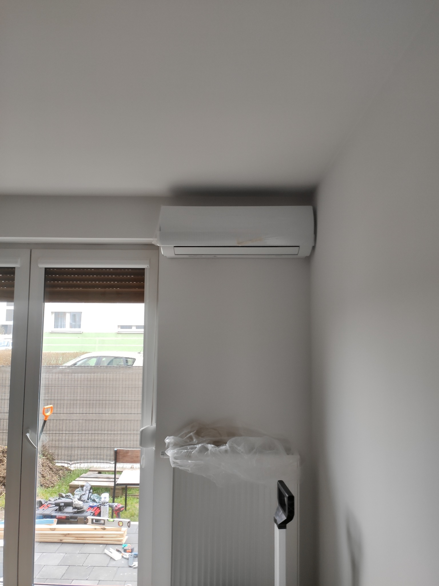 Zamontowany klimatyzator na ścianie w jasnym pomieszczeniu, widoczne narzędzia budowlane i materiały przez otwarte okno.