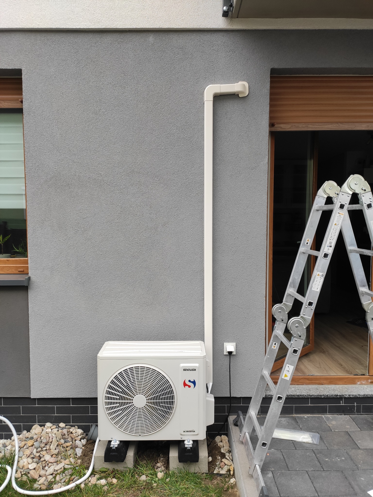 Zewnętrzna jednostka klimatyzacji Sinclair zamontowana na betonowych podstawach przy szarej ścianie domu z widoczną białą rurą odprowadzającą, obok rozstawiona aluminiowa drabina.