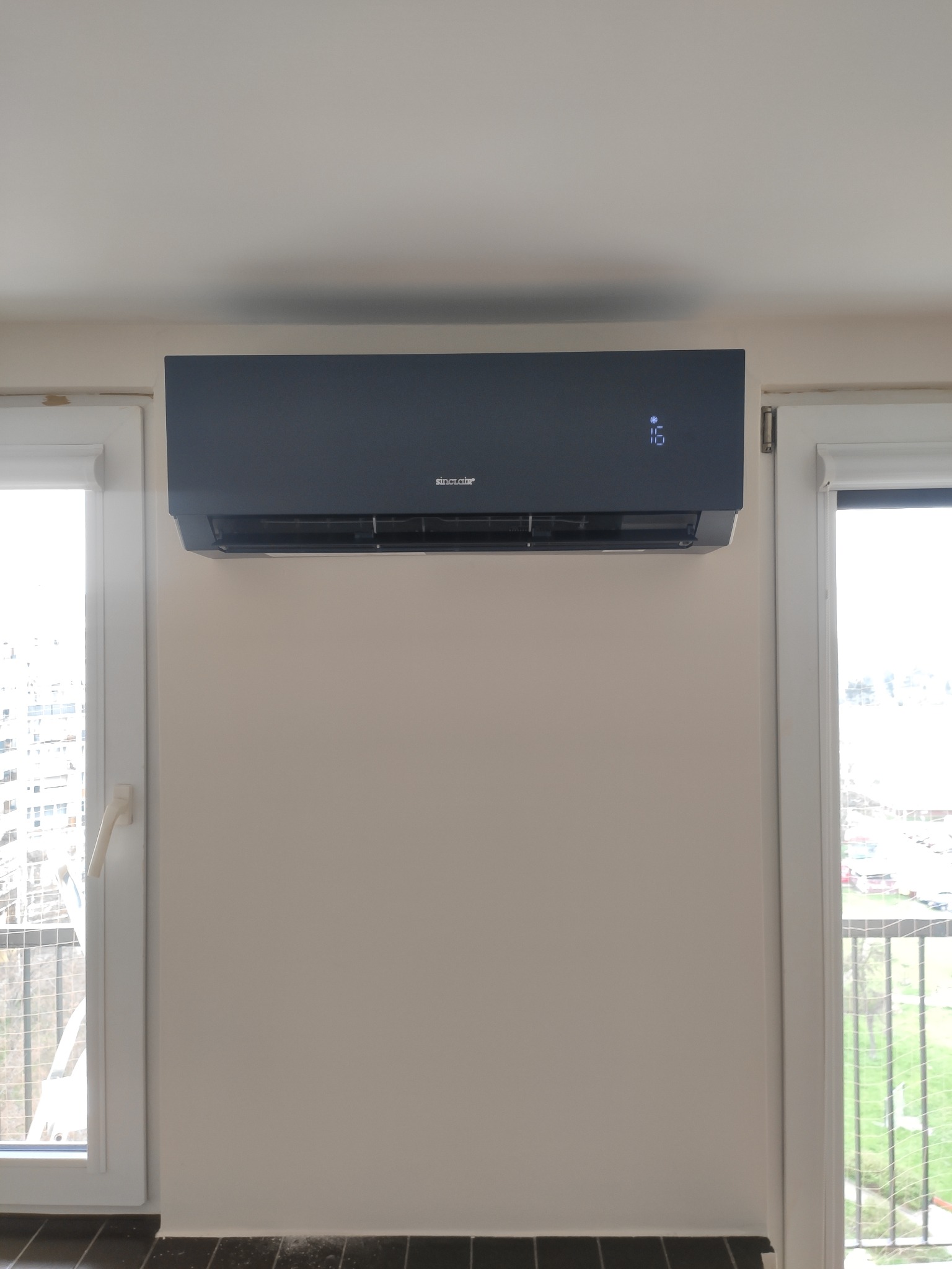 Czarny klimatyzator Sincooler zamontowany na ścianie nad oknem z widokiem na miasto, ustawiony na 16 stopni.