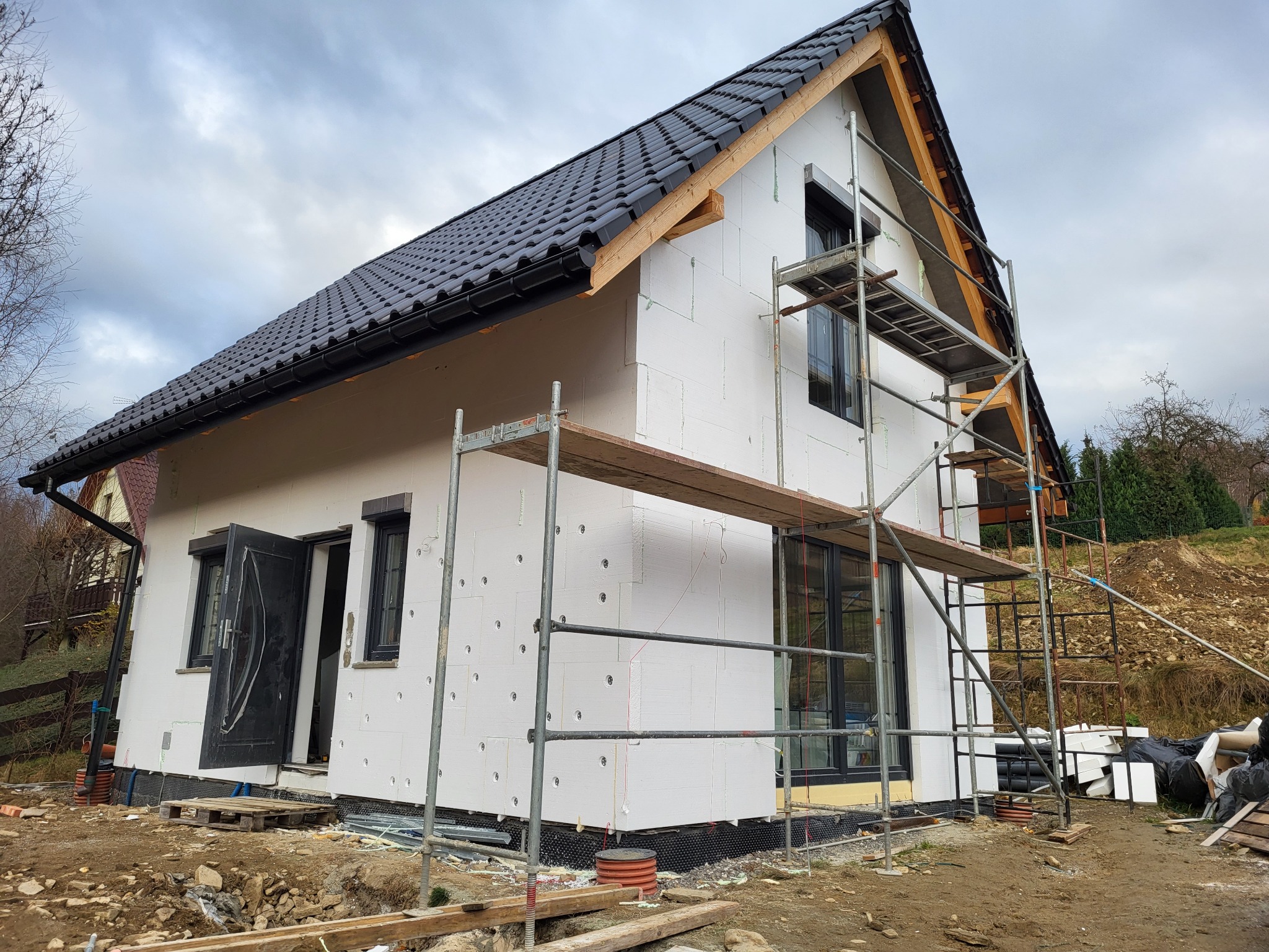 Nowoczesny dom jednorodzinny w trakcie ocieplania elewacji styropianem, widoczne rusztowania i elementy konstrukcyjne dachu przed montażem podbitki.
