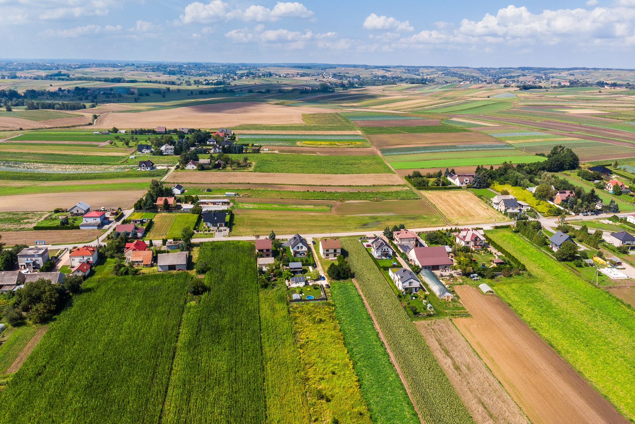 Panoramiczny widok z lotu ptaka na wieś z domami jednorodzinnymi, polami uprawnymi i zielenią, w słoneczny dzień z lekkimi chmurami.