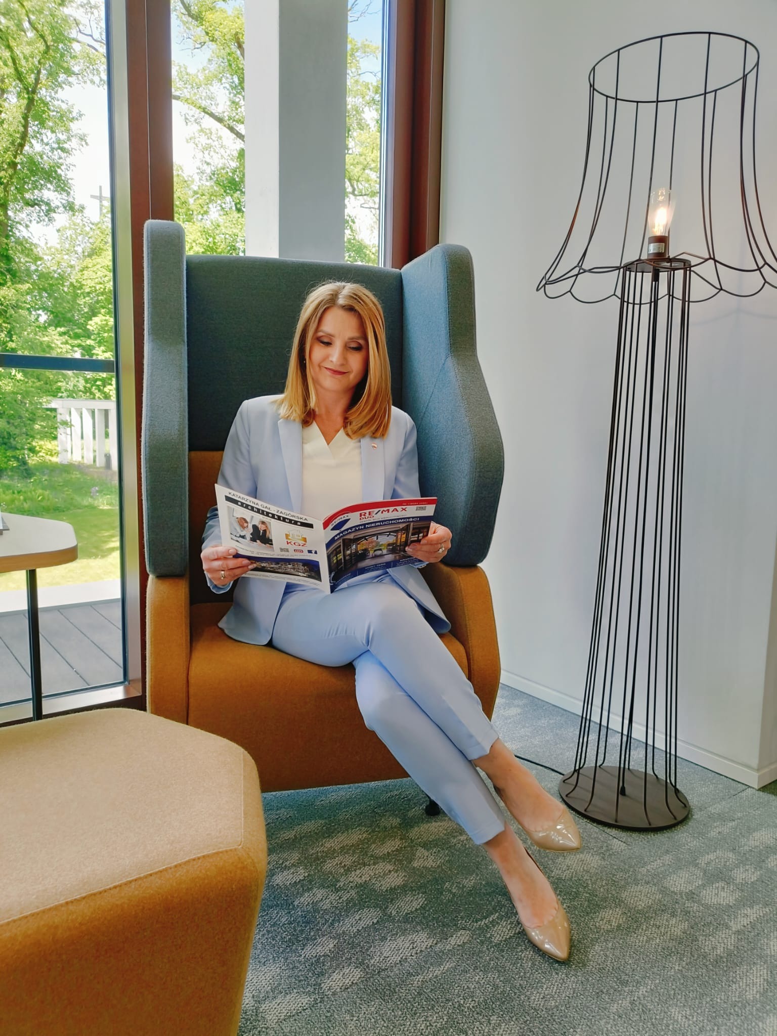 Kobieta w błękitnym garniturze siedzi w fotelu uszak, czytając magazyn nieruchomości z logo RE/MAX, obok stoi lampa z metalowym ażurowym kloszem, widok z okna na zieleń.