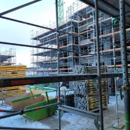 Budowa wielopiętrowego budynku w trakcie realizacji, widoczne rusztowania, dźwig i materiały budowlane składowane na placu budowy