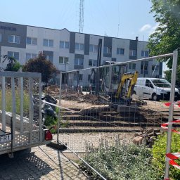 Kamil Dutkiewicz - Żółta mini koparka przy budynku Policji, otoczona metalowym ogrodzeniem i taśmą ostrzegawczą. Widoczny wykop, przyczepa i częściowo zasłonięty biały samochód.