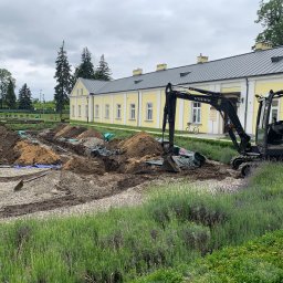 Kamil Dutkiewicz - Czarny mini-koparka Volvo EC35C na tle żółtego budynku. W tle wykopane rowy, kamienie i ziemia. Prace ziemne w toku, widok na budynek i zieleń.