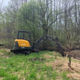 Kamil Dutkiewicz - Żółta mini koparka Volvo EC35C w zielonym terenie z drzewami w tle, wykonująca prace ziemne przy cieku wodnym. Widoczne ślady opon.