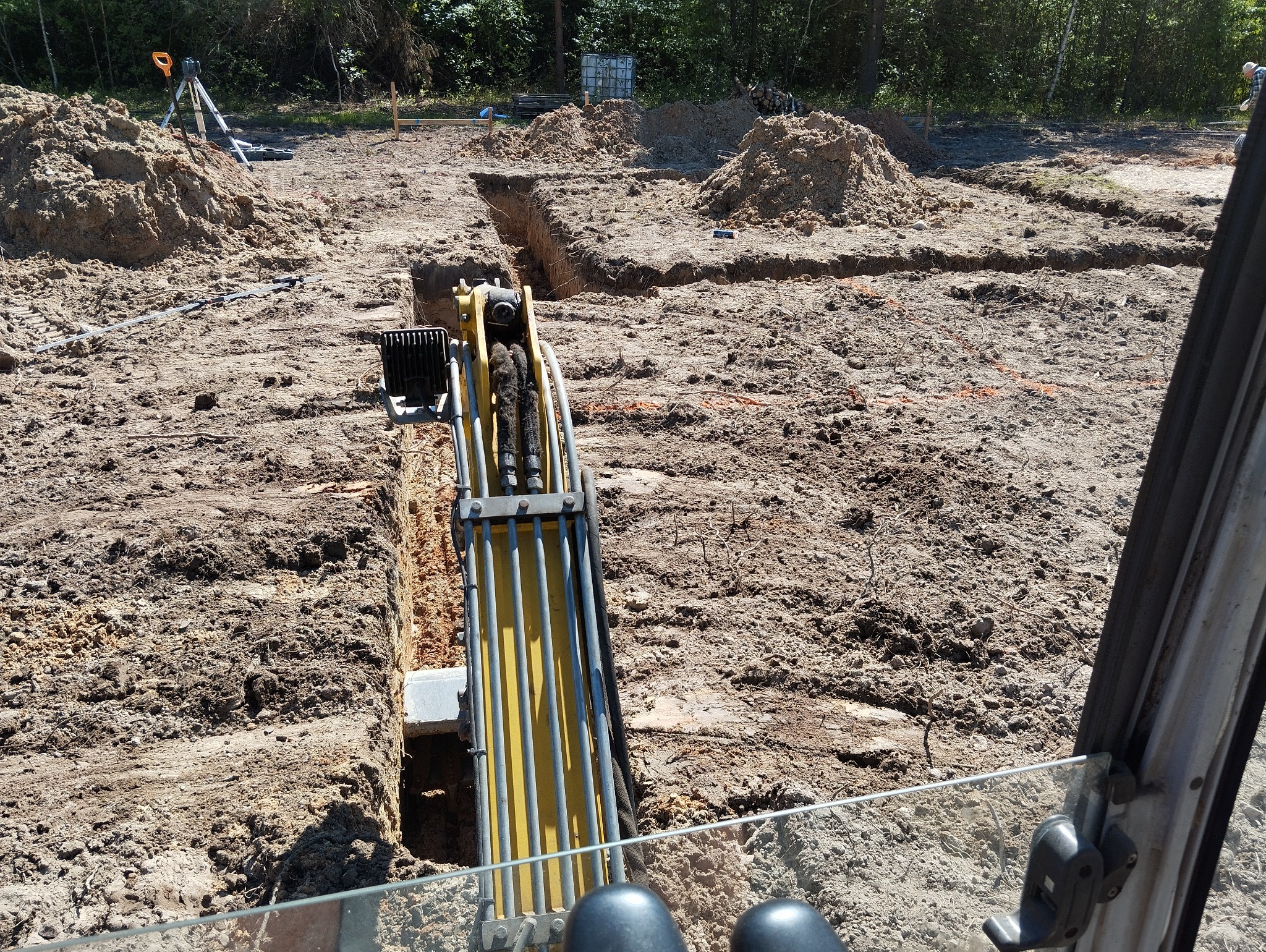 Widok z kabiny koparki na wykop pod fundamenty, widoczne pryzmy ziemi, niwelator i wytyczone linie wykopu.