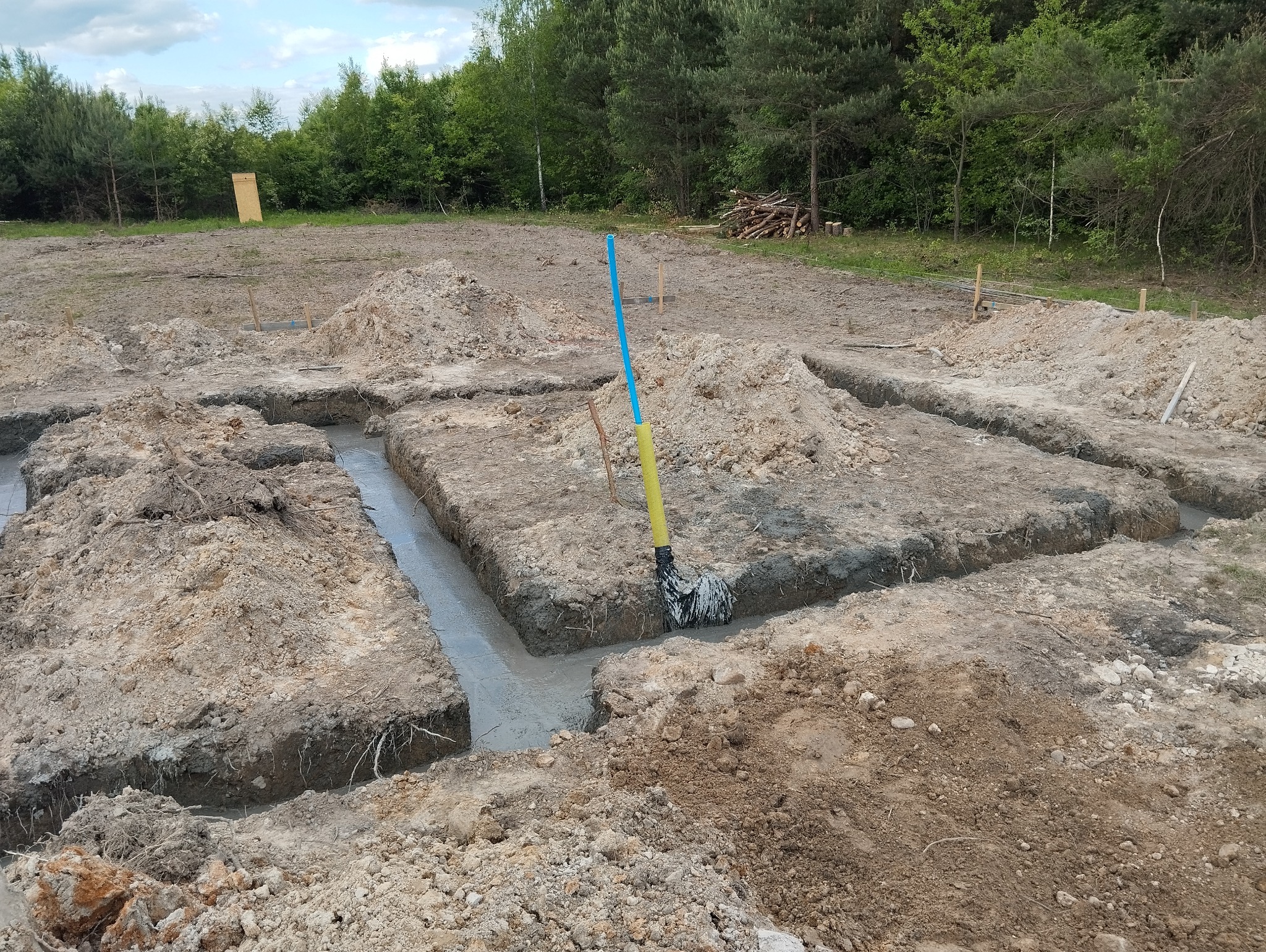 Wykopane rowy pod fundamenty wypełnione świeżym betonem, widoczne drewniane pale wyznaczające granice, w tle las i stos drewna.
