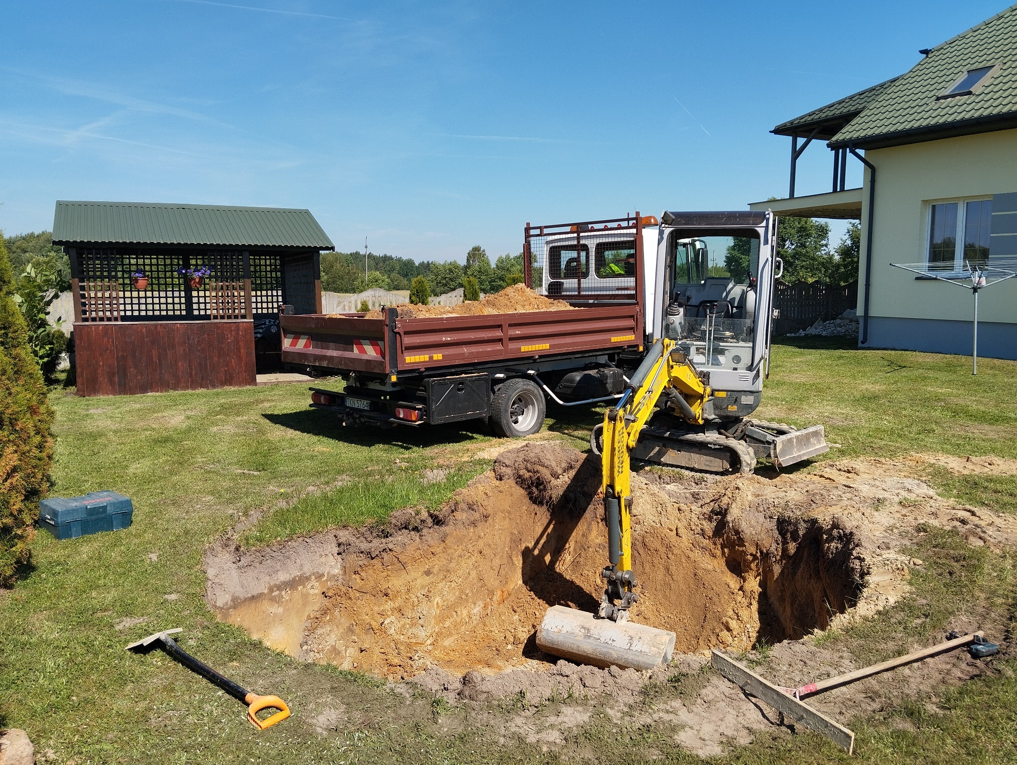 Żółta mini koparka Wacker Neuson przy wykopie w ziemi, obok ciężarówka z ziemią, na trawie leżą narzędzia, w tle dom i altana.