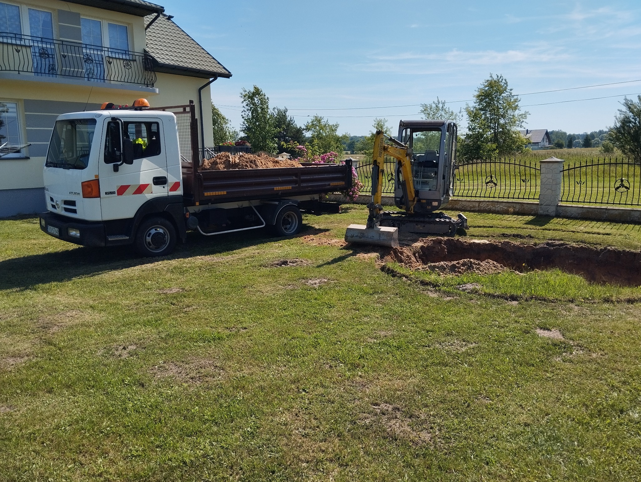 Żółta minikoparka przy wykopie w trawniku, obok biała ciężarówka z ziemią, dom w tle