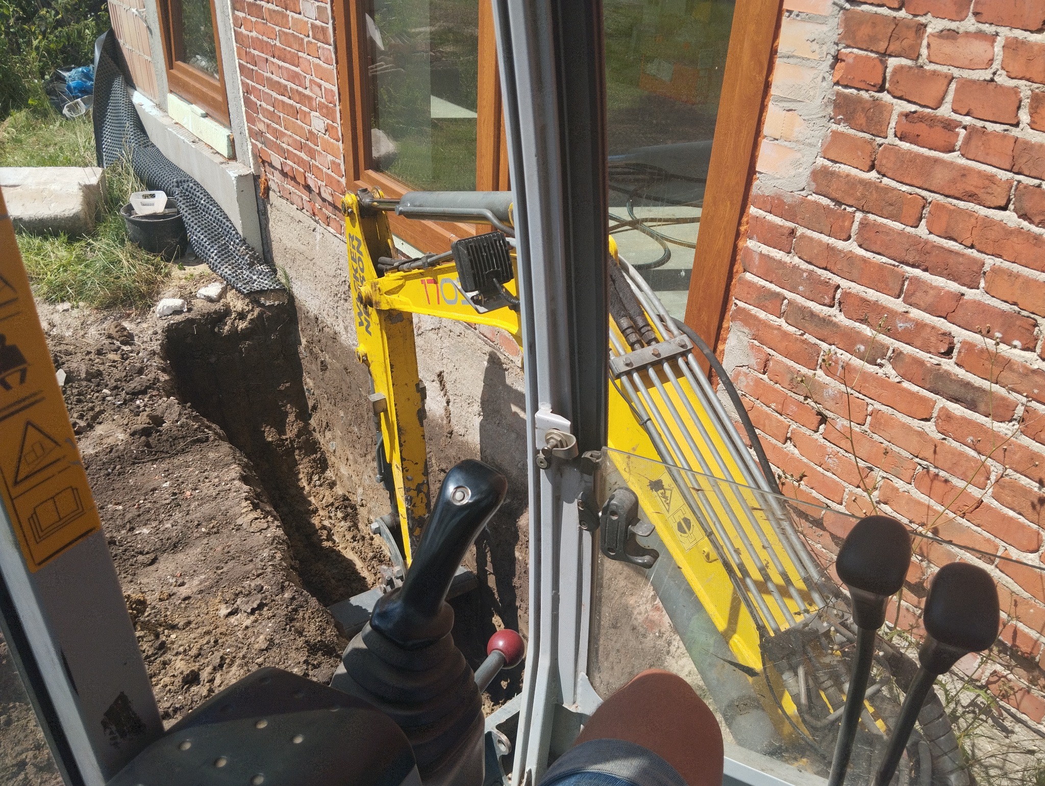 Widok z kabiny mini koparki podczas wykonywania wąskiego wykopu wzdłuż fundamentu budynku z czerwonej cegły, widoczna izolacja fundamentu i drenaż.