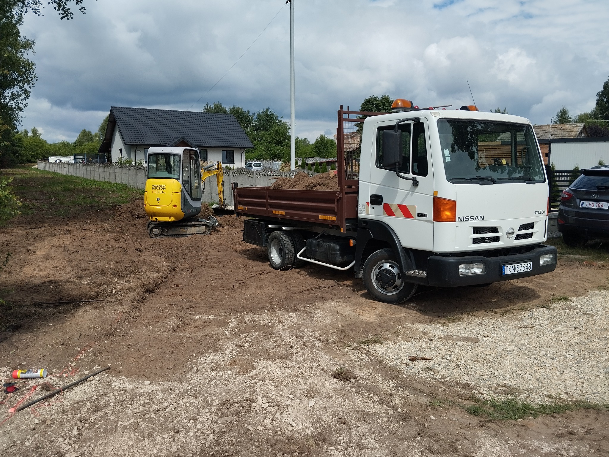 Żółta mini koparka Wacker Neuson 1703 i biała ciężarówka Nissan z brązową skrzynią ładunkową wypełnioną ziemią na placu budowy, w tle dom z szarym dachem i betonowe ogrodzenie.