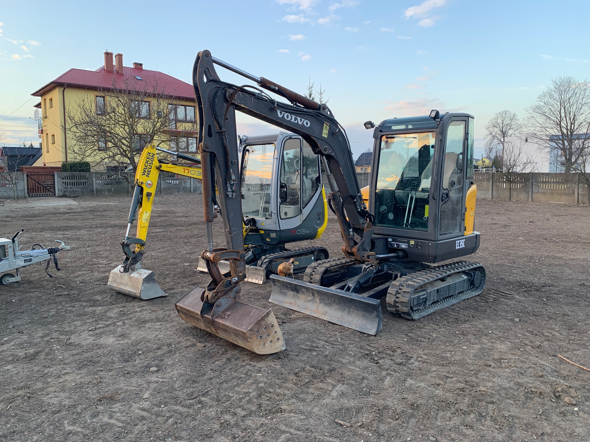 Dwa mini-koparki Volvo EC35C i Wacker Neuson 1703 na placu budowy, widoczne ślady gąsienic w ziemi, w tle budynek mieszkalny z czerwonym dachem i ogrodzenie betonowe.