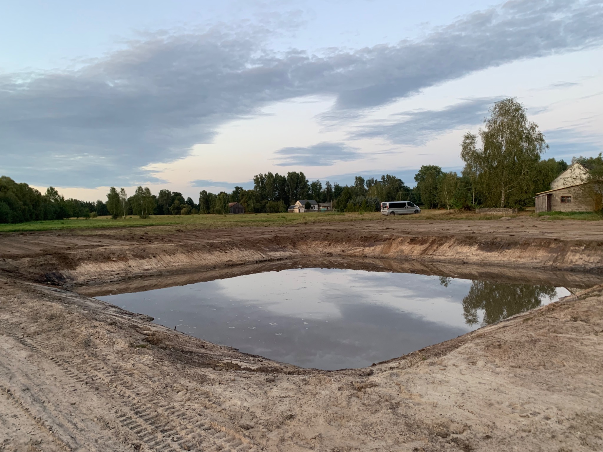 Nowo wykopany, wypełniony wodą staw o regularnym kształcie na tle krajobrazu z drzewami i budynkami. Ślady opon na ziemi. Pochmurne niebo.