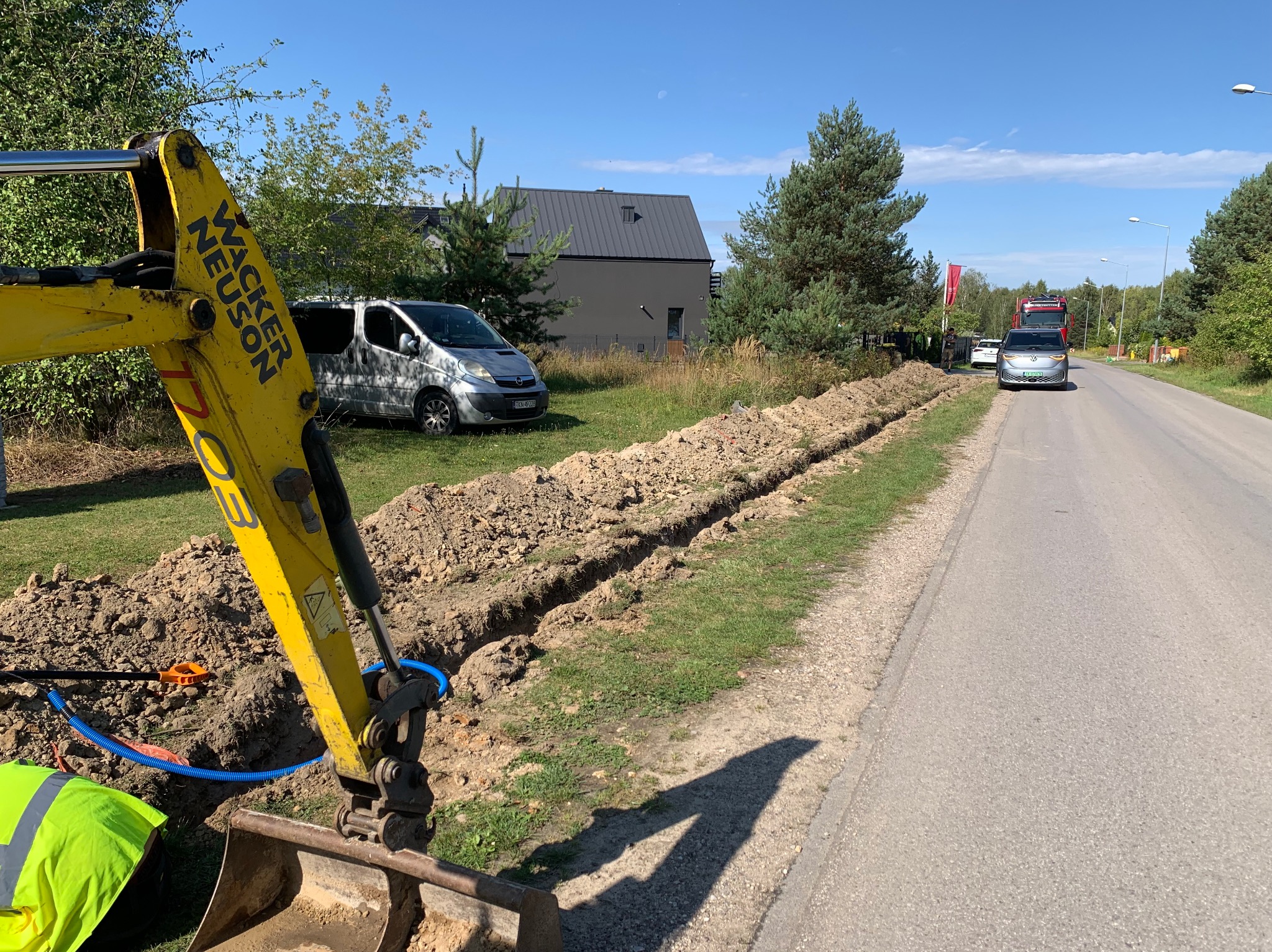 Żółta koparka Wacker Neuson na tle wykopu wzdłuż drogi asfaltowej, widoczne rury instalacyjne i budynek mieszkalny w oddali. Dzień słoneczny.