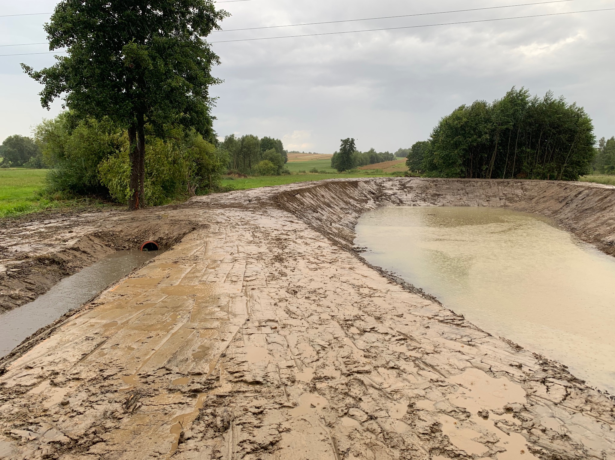 Świeżo wykopany, ziemny staw w krajobrazie wiejskim, z widocznymi śladami opon w błotnistej ziemi i częściowo wypełniony wodą, pod pochmurnym niebem.