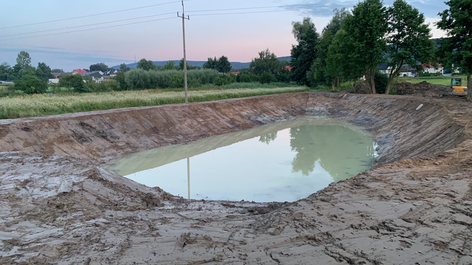 Wykopany staw wypełniony wodą, ziemne brzegi, w tle zieleń i zabudowania. Widoczny fragment koparki. Zdjęcie wykonane o zmierzchu, pochmurne niebo.