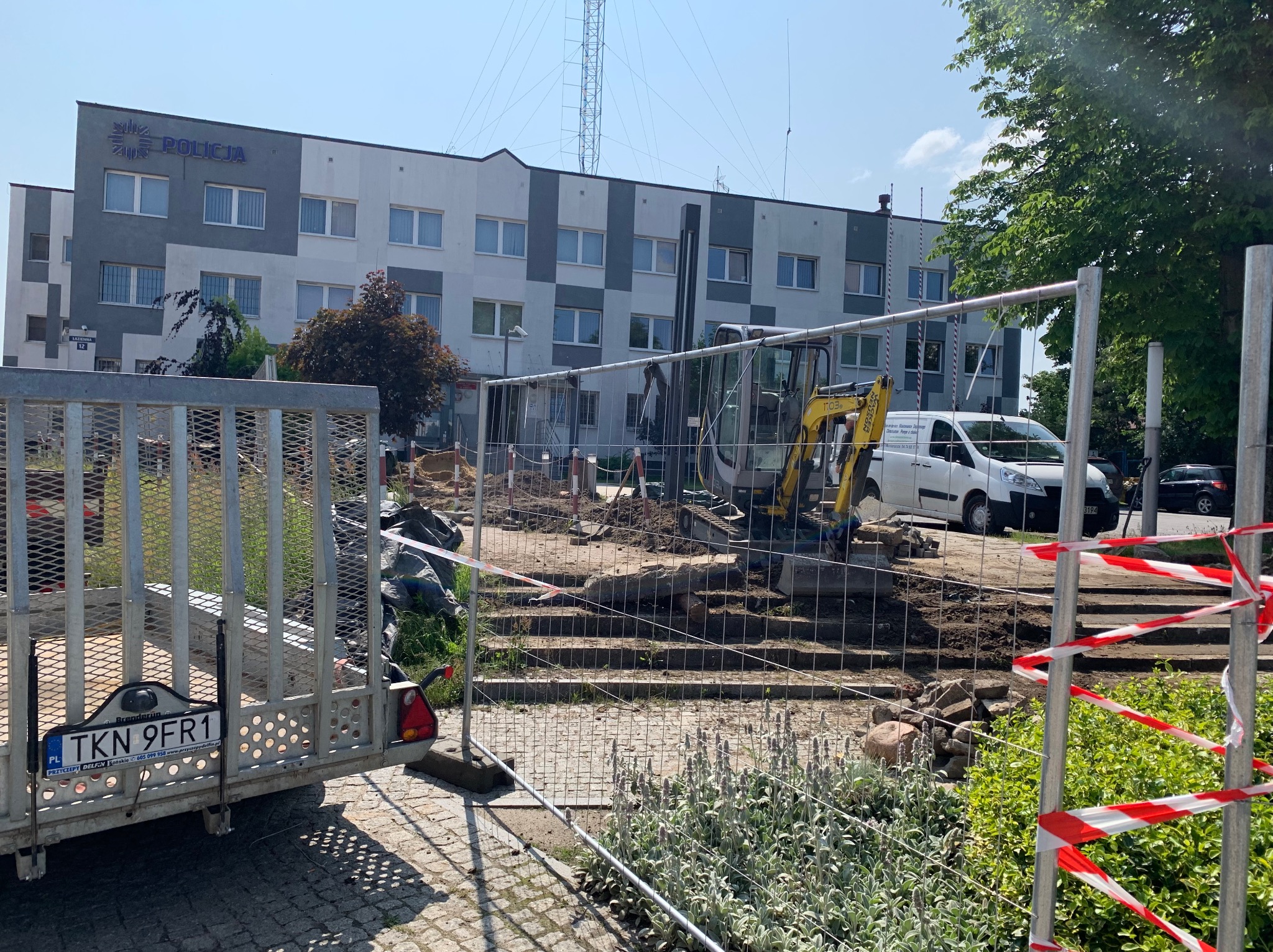 Żółta mini koparka przy budynku Policji, otoczona metalowym ogrodzeniem i taśmą ostrzegawczą. Widoczny wykop, przyczepa i częściowo zasłonięty biały samochód.