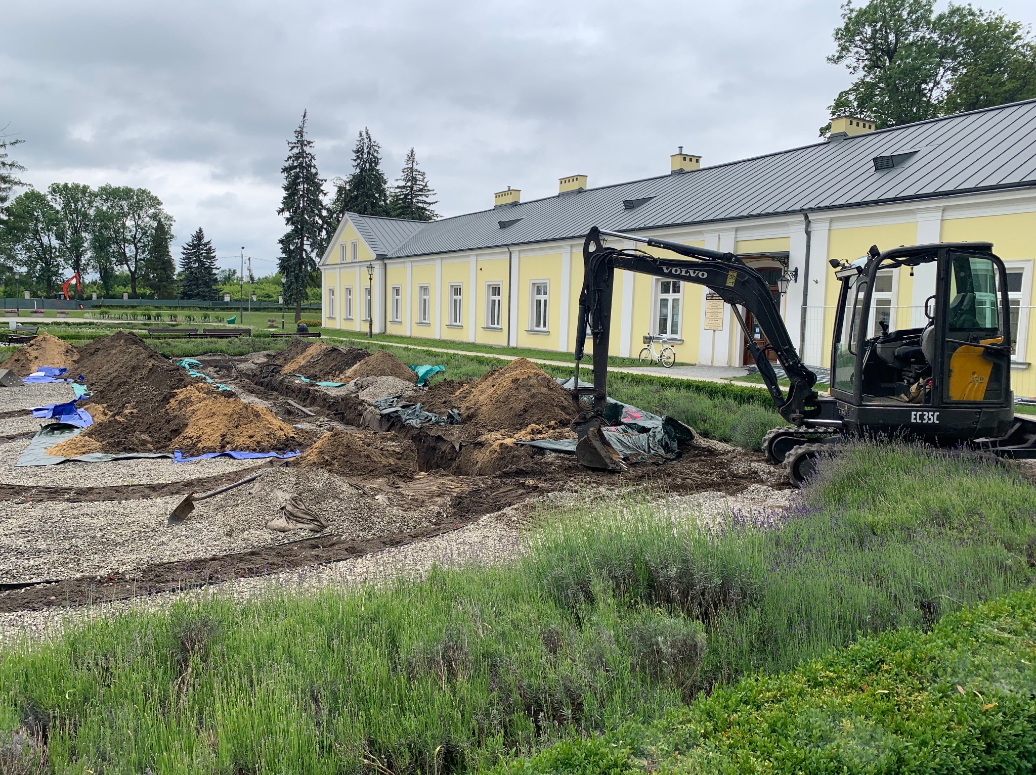 Czarny mini-koparka Volvo EC35C na tle żółtego budynku. W tle wykopane rowy, kamienie i ziemia. Prace ziemne w toku, widok na budynek i zieleń.