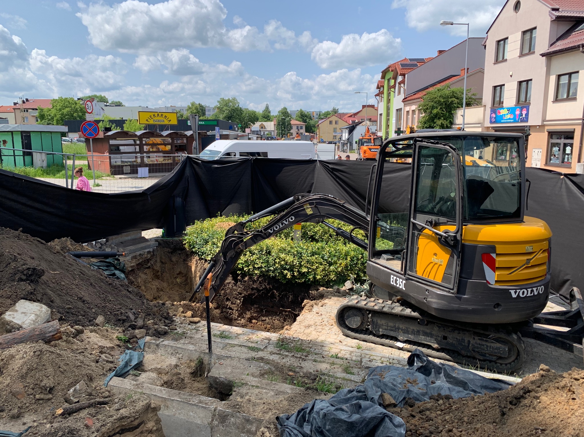 Żółta koparka Volvo EC35C w trakcie prac ziemnych na ulicy miejskiej, wykop zabezpieczony czarną folią, w tle zabudowa miejska i błękitne niebo.