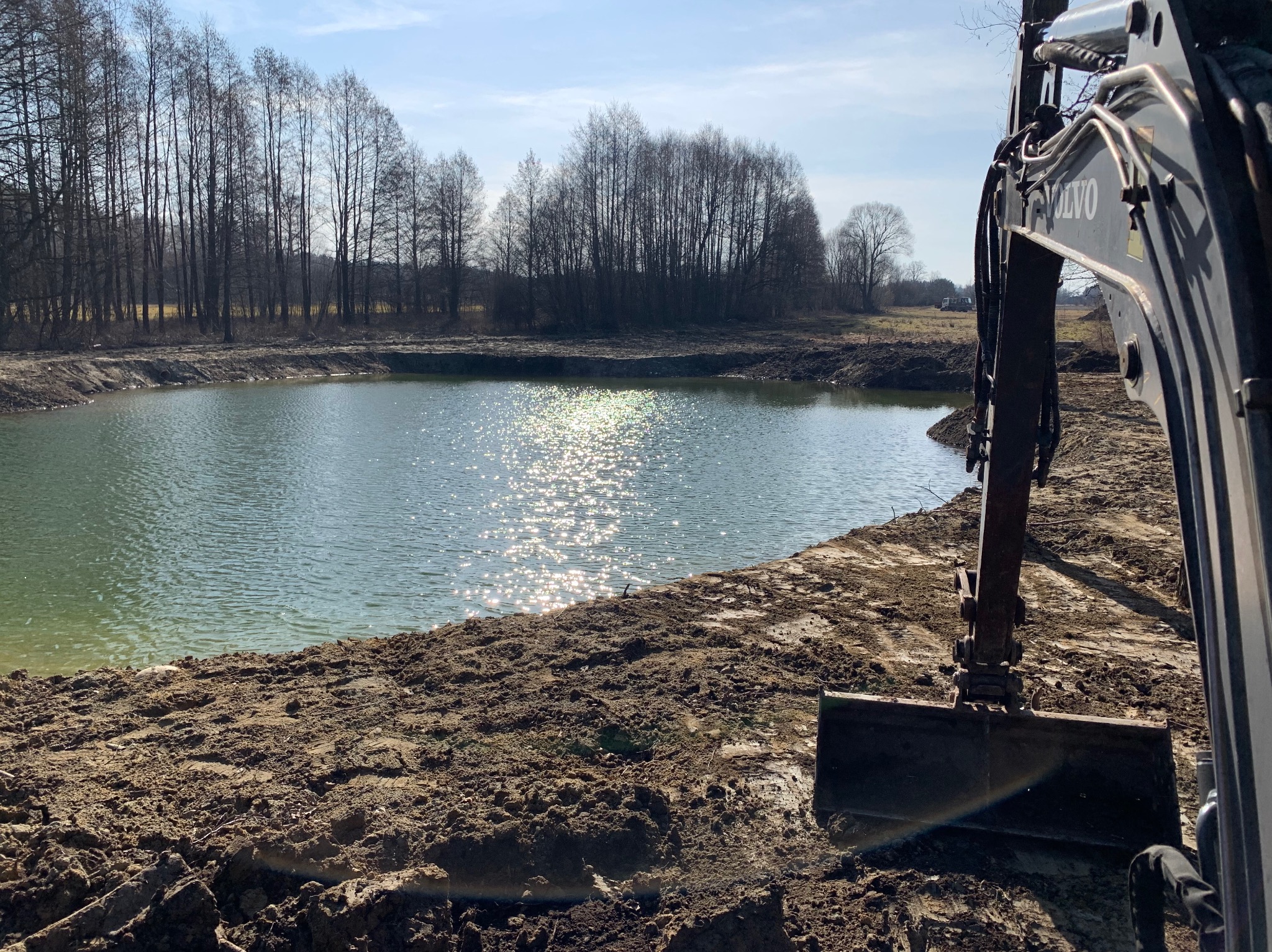 Wykopany staw w słoneczny dzień. Brzeg ziemny, woda z refleksami. Na pierwszym planie ramię koparki Volvo. W tle drzewa bez liści i błękitne niebo.